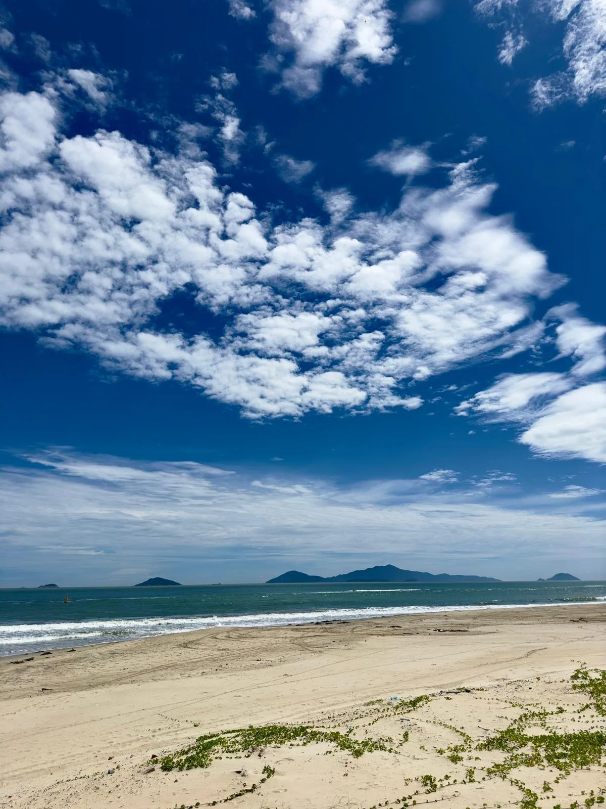 Uitzicht op een zandstrand met de zee en kleine eilandjes op de achtergrond onder een bewolkte blauwe hemel.