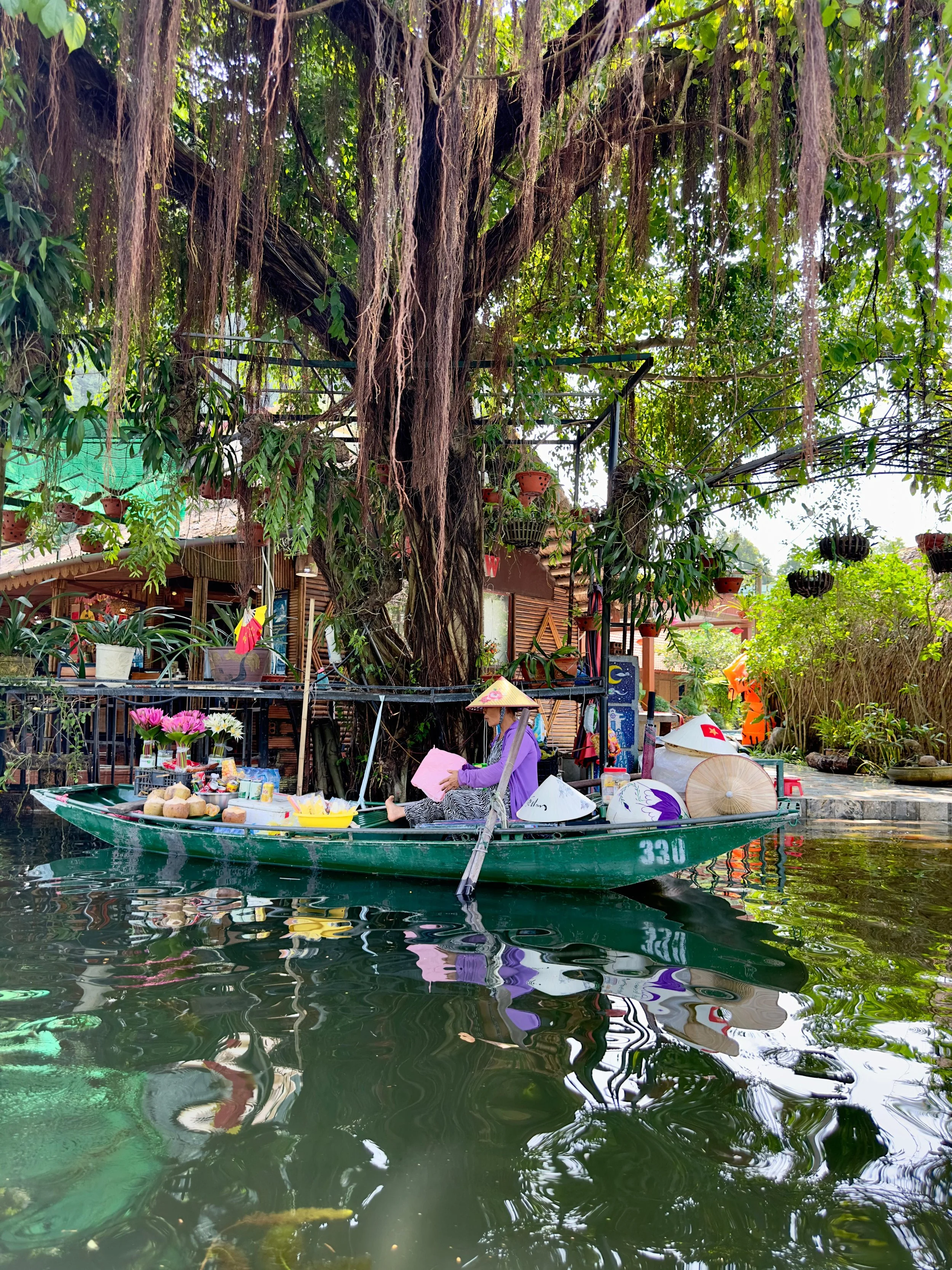 Vrouw in traditionele Vietnamese kleding op een roeiboot vol bloemen en fruit, varend op een kalme rivier met een groot boom en groene planten op de achtergrond.