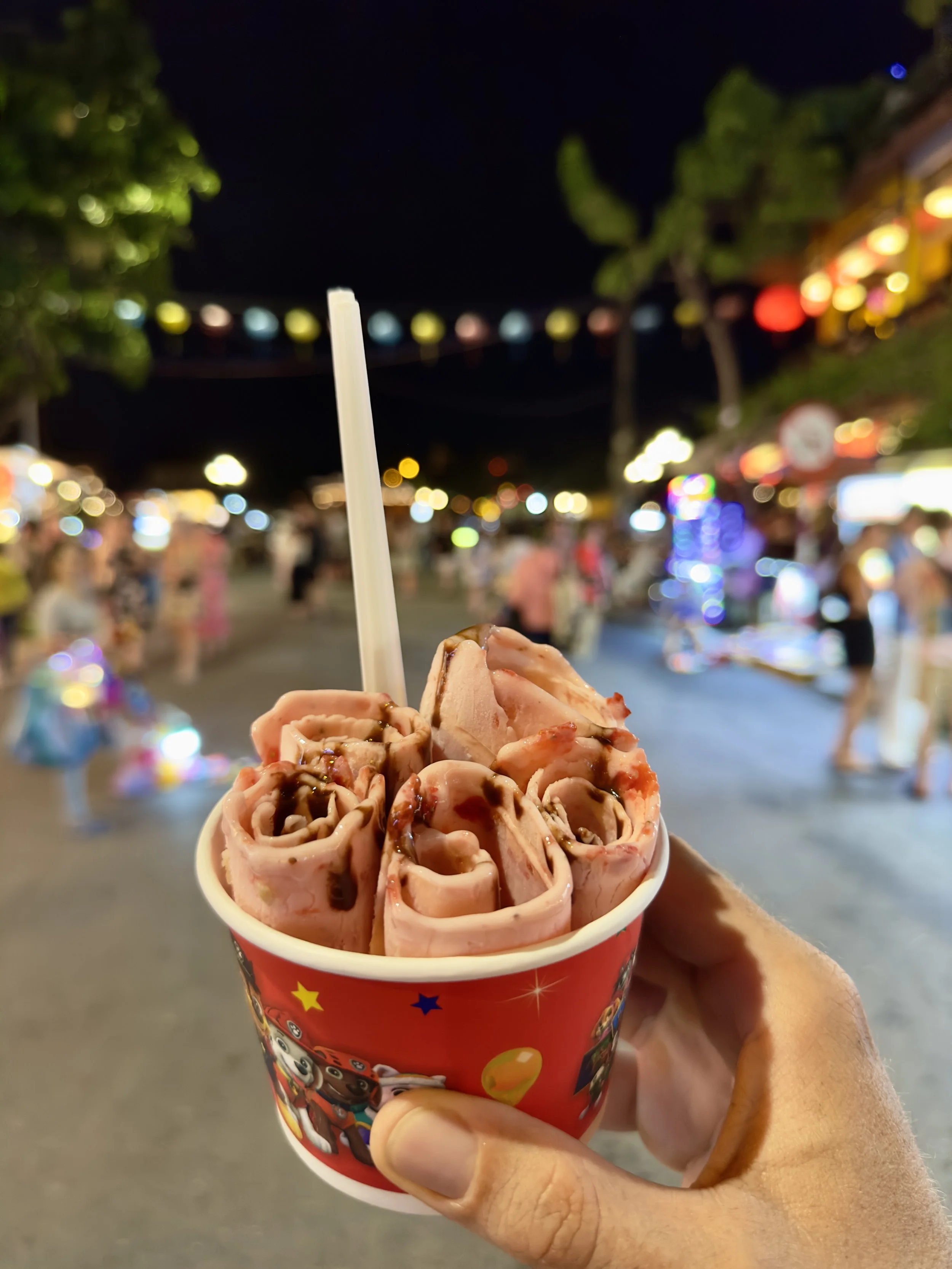 Een hand die een rood cupje met roze ijs met chocolade en andere saus drinkt, op een levendige markt 's avonds met kleurrijke lichten en mensen op de achtergrond.