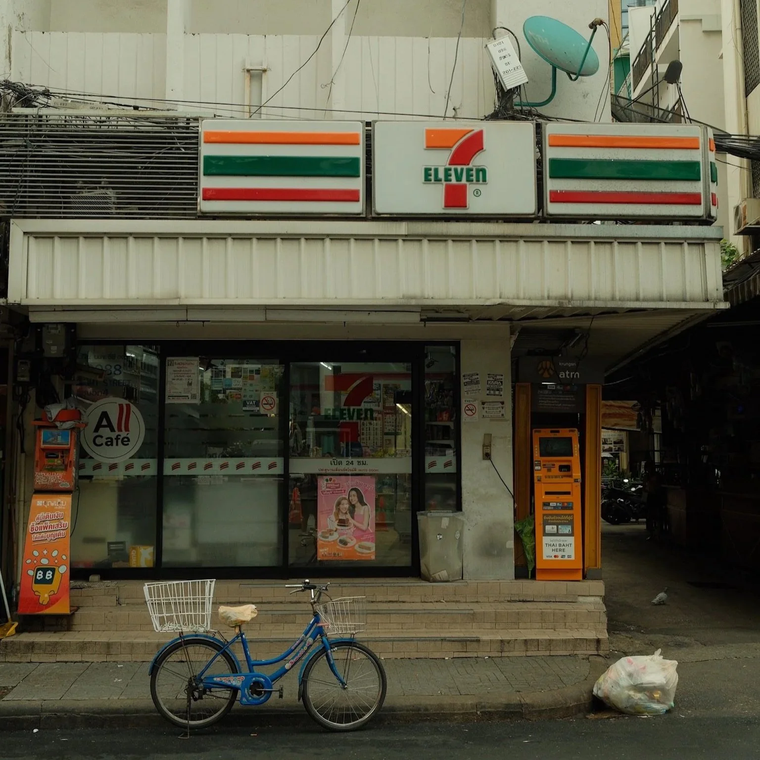 Een supermarkt met het logo van 7-Eleven, een blauwe fiets met manden voor en achter geparkeerd voor de ingang, en een vuilniszak op de stoep.