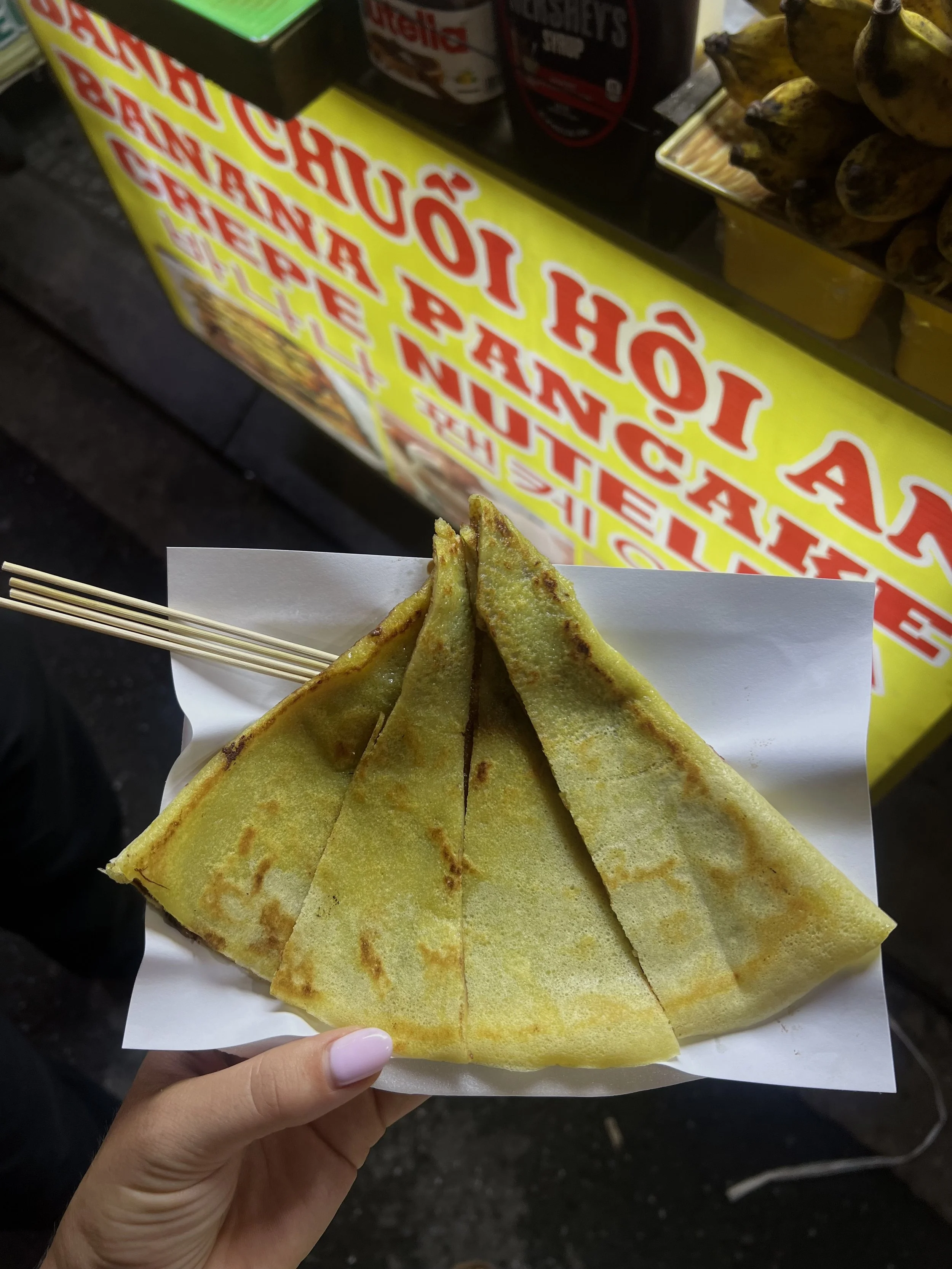 Tienkante bananenpannekoek op een papieren bord met houten satéprikkers, handen erachter in een straatvoedselstand.