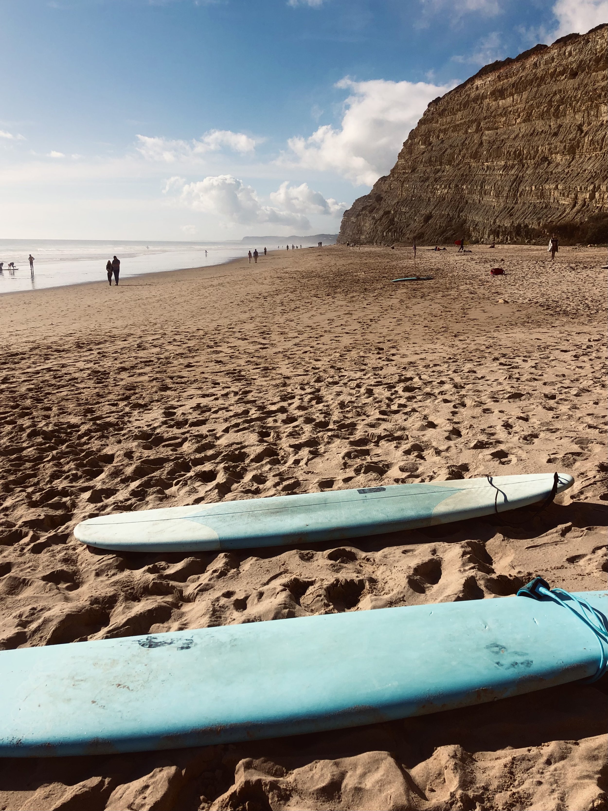 Tienersurfborden op zandstrand met kliffen op de achtergrond, zee en paar wandelaars, bewolkte hemel.