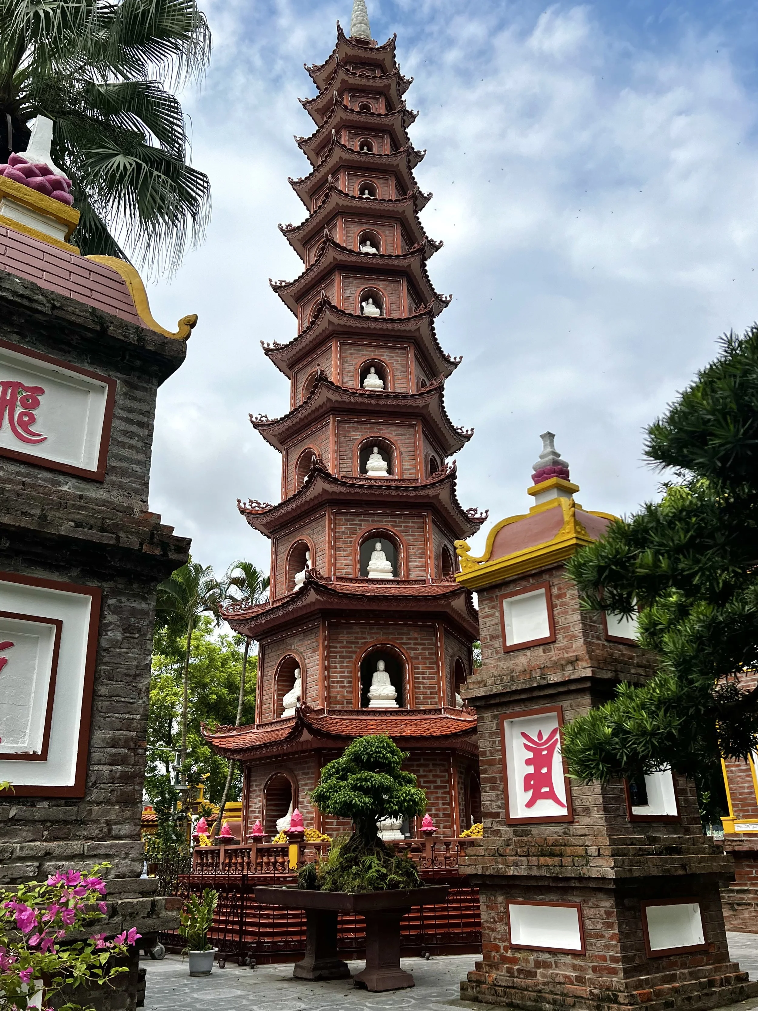Een Chinese pagode met meerdere verdiepingen, gemaakt van rood baksteen, omgeven door bomen en bloemen, onder een bewolkte hemel.