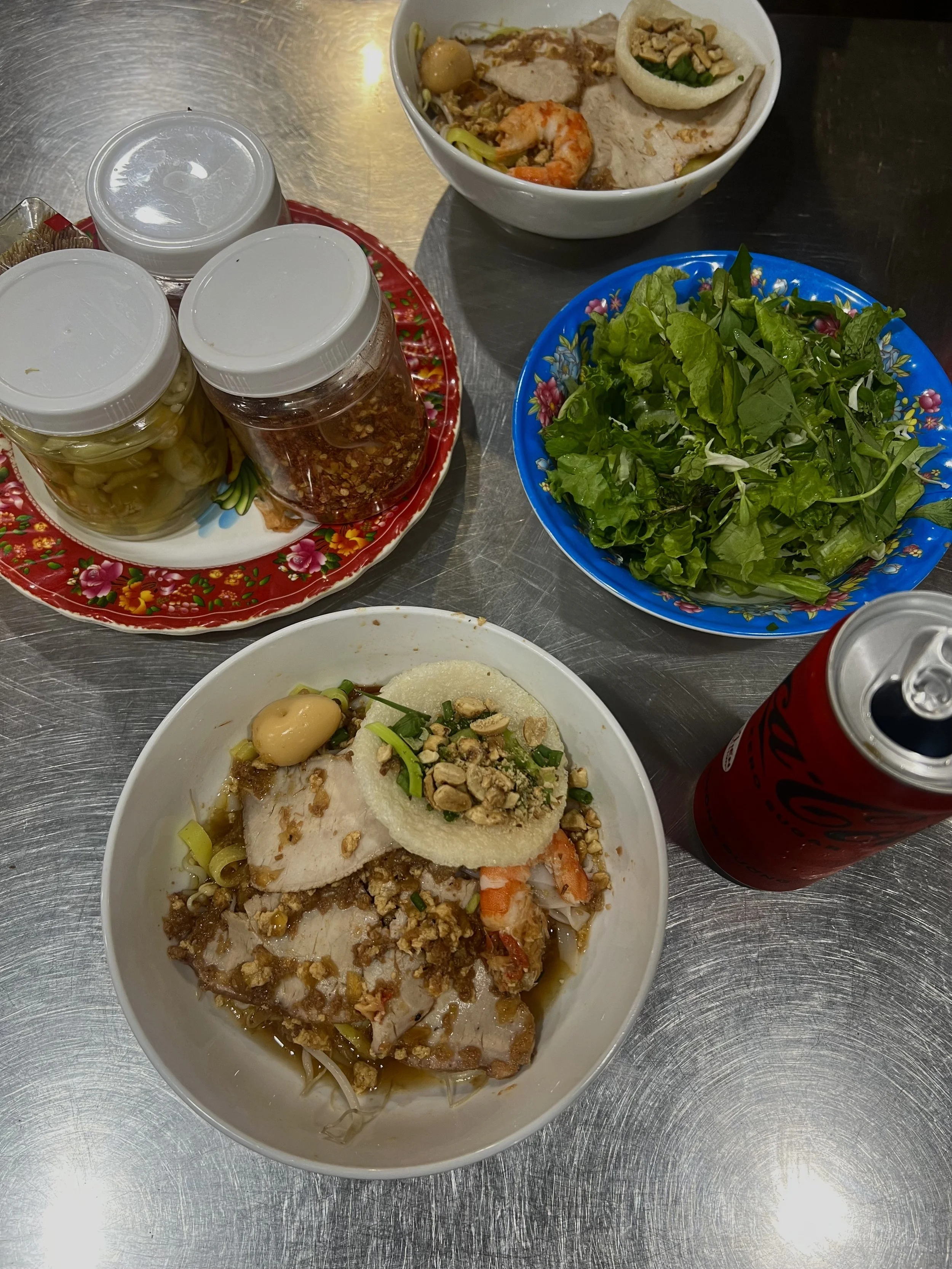Streetfood eten met een bowl met noodles, groenten, ei. Op een ijzeren tafel waar verschillende sauzen en cola op staan.