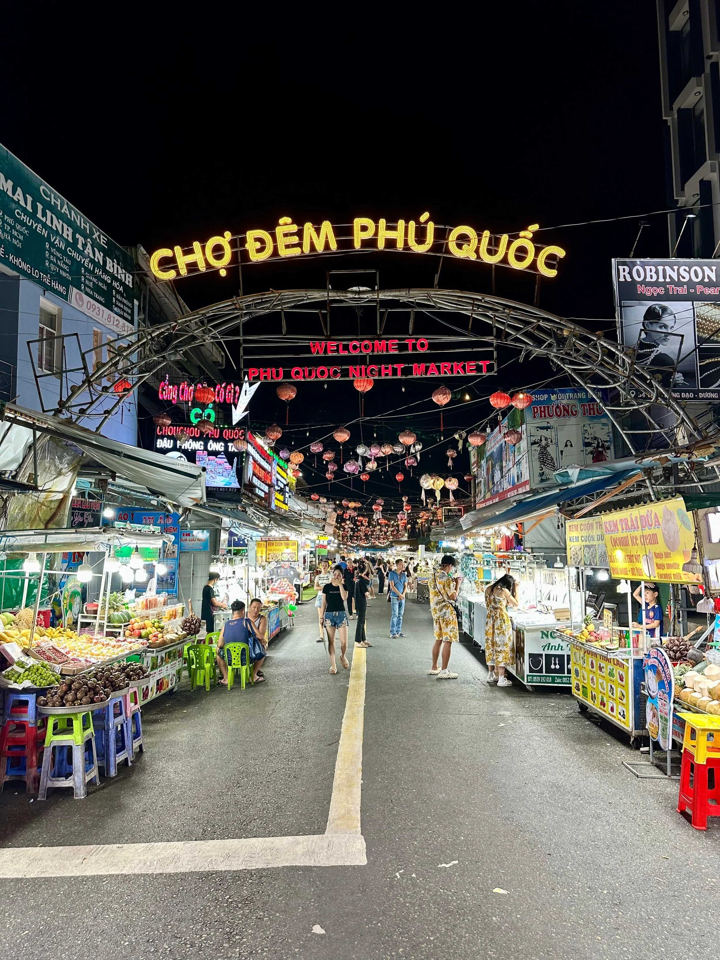 Openluchtmarkt met straatverkopers, fruit en andere etenswaren, verlichte hangende decoraties, en neonborden, 'Chợ Đêm Phú Quốc' in gele letters boven de ingang.