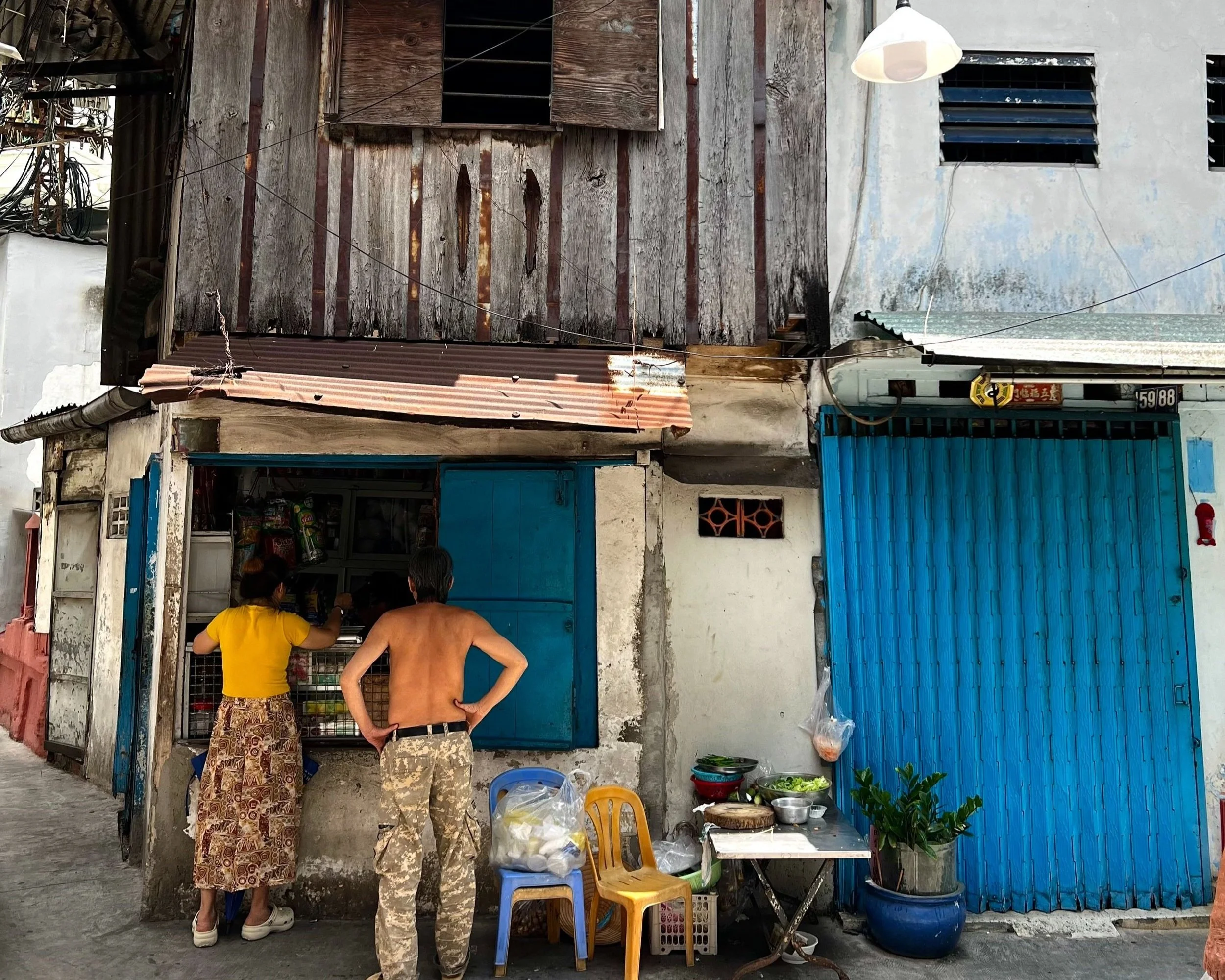 Twee mensen bij een kleine straatwinkel, een vrouw in een gele blouse en een lange rokje, en een man zonder shirt met camouflagebroek, kopen eten of drinken. Er staan plastic stoelen, een tafel met groenten en een plant ernaast.