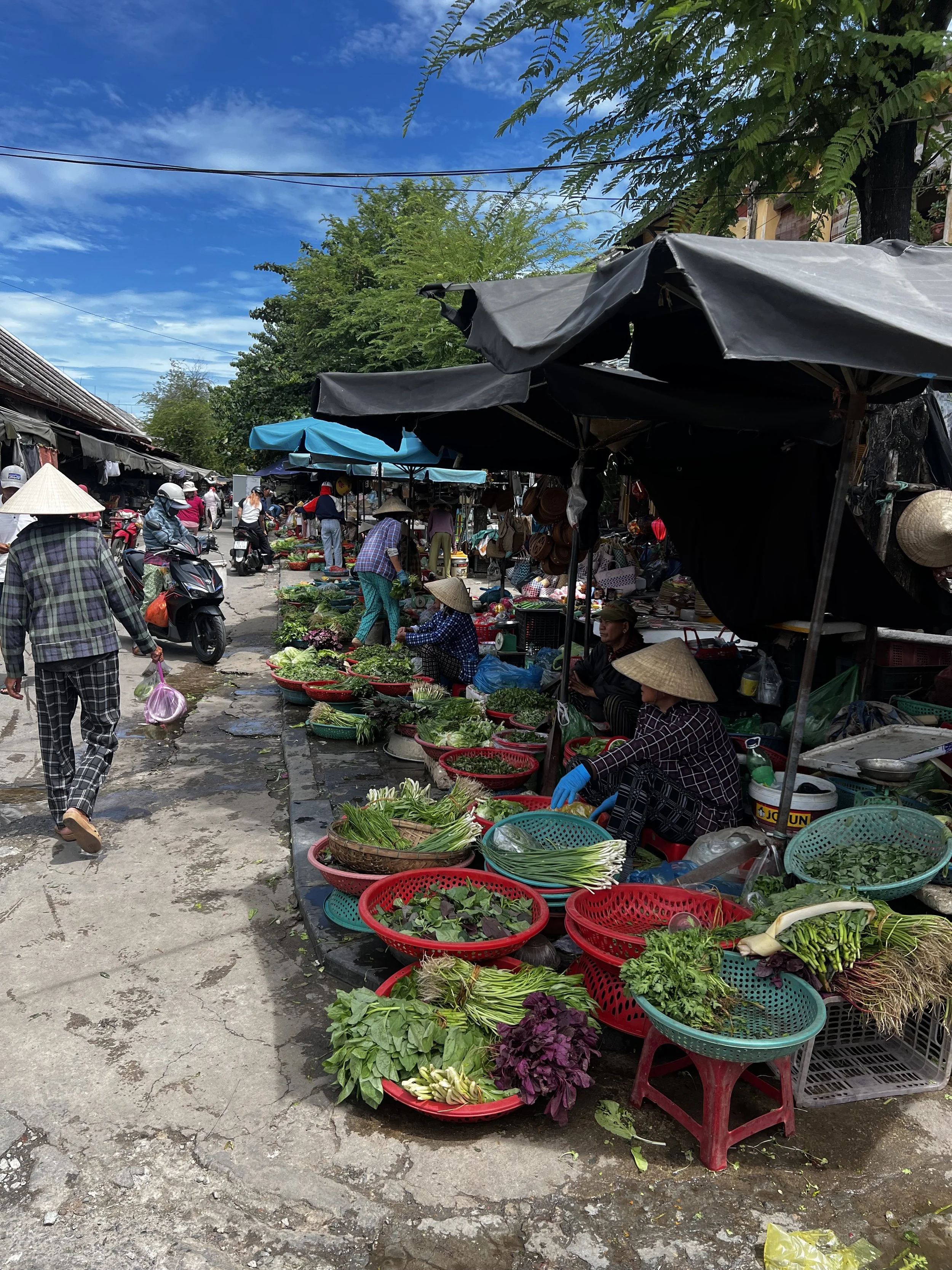 Openluchtmarkt met groenten en fruit verkocht door lokale verkopers onder parasols, mensen lopen rond in een stad op een heldere dag.