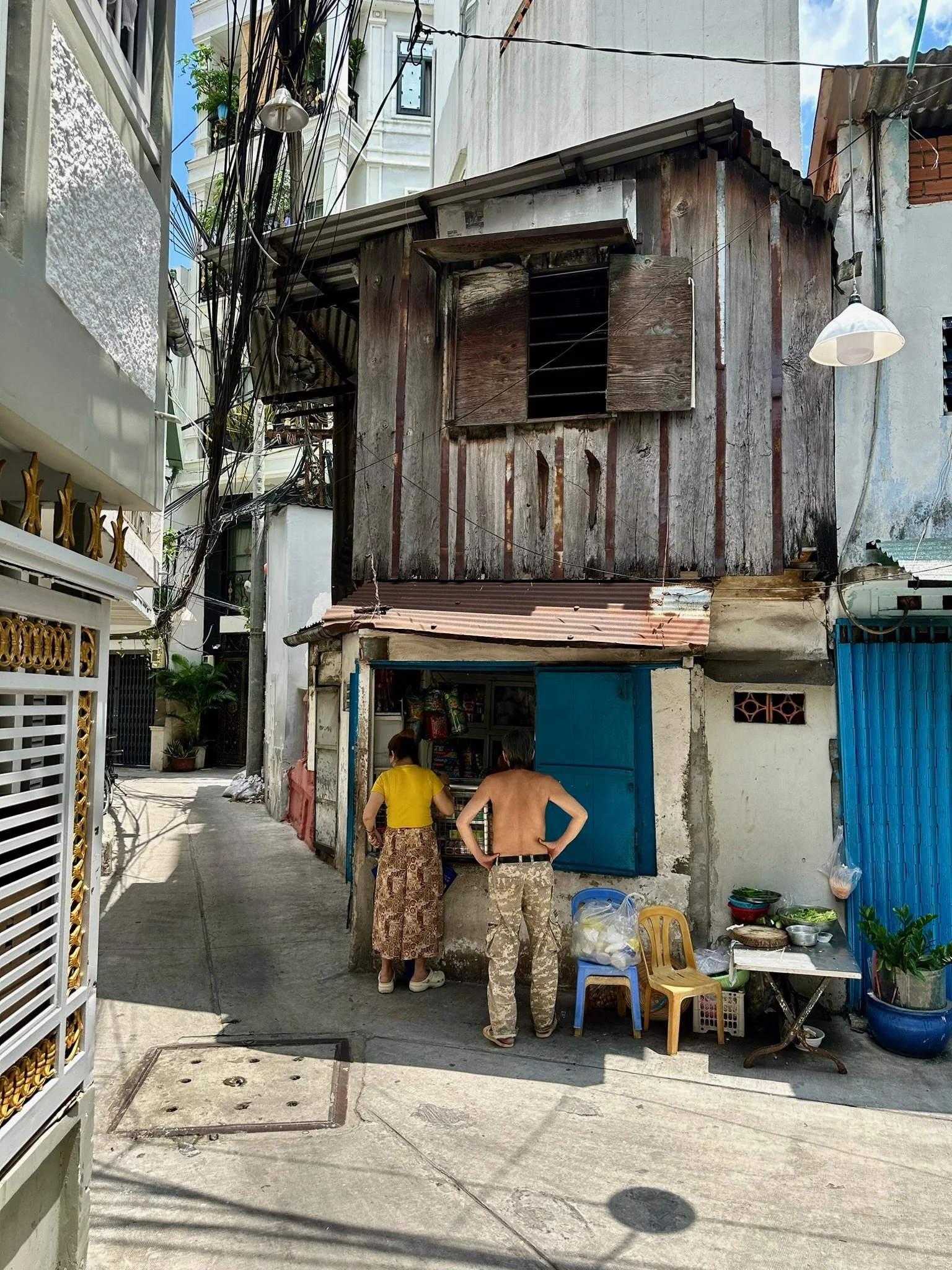 Twee mensen staan voor een kleine winkel in een steegje, een vrouw met een gele blouse en een lange rok, en een shirtloze man in camouflages. De winkel heeft een blauwe deur en is gevestigd in een oud gebouw met houten gevel en kapotte luiken. Buiten staan plastic stoelen en een kleine tafel met groenten en andere producten.