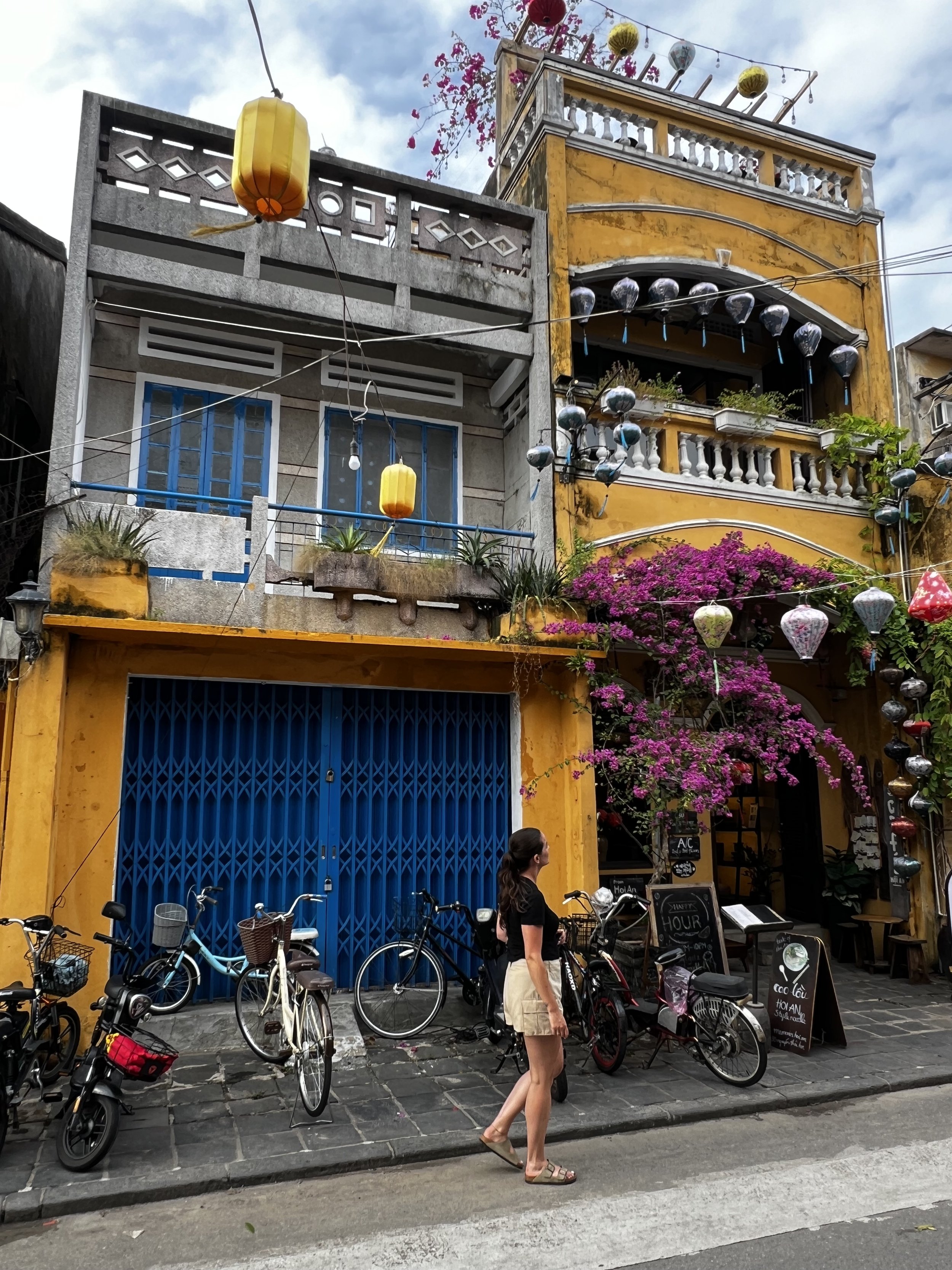 Kleurrijke straat met fietsen, een vrouw, felgele en blauwe gevels, bloemen, en hanging lampions.