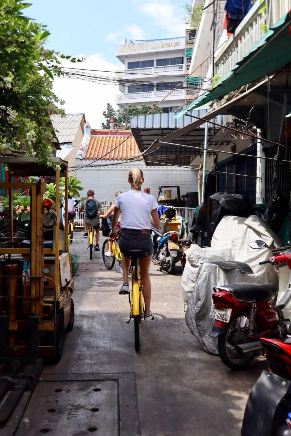 Mensen fietsen door een smalle straat in een stadswijk, omgeven door gebouwen, scooters en fietsen.