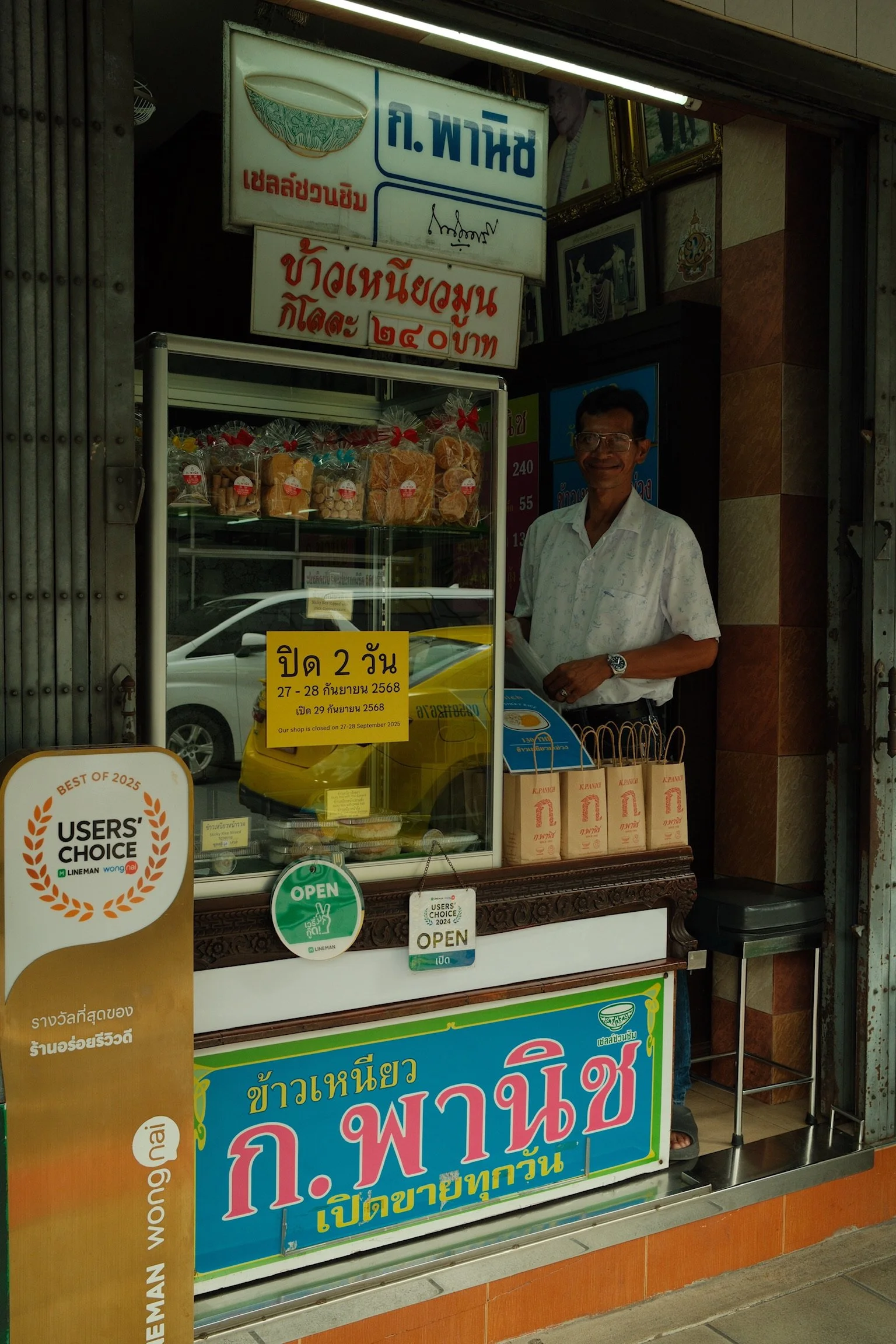 Kleine snackkraam met een man achter de toonbank, kleurrijke verpakkingen, en borden met Thaise tekst en prijzen. Een bord geeft aan dat de winkel twee dagen gesloten is.