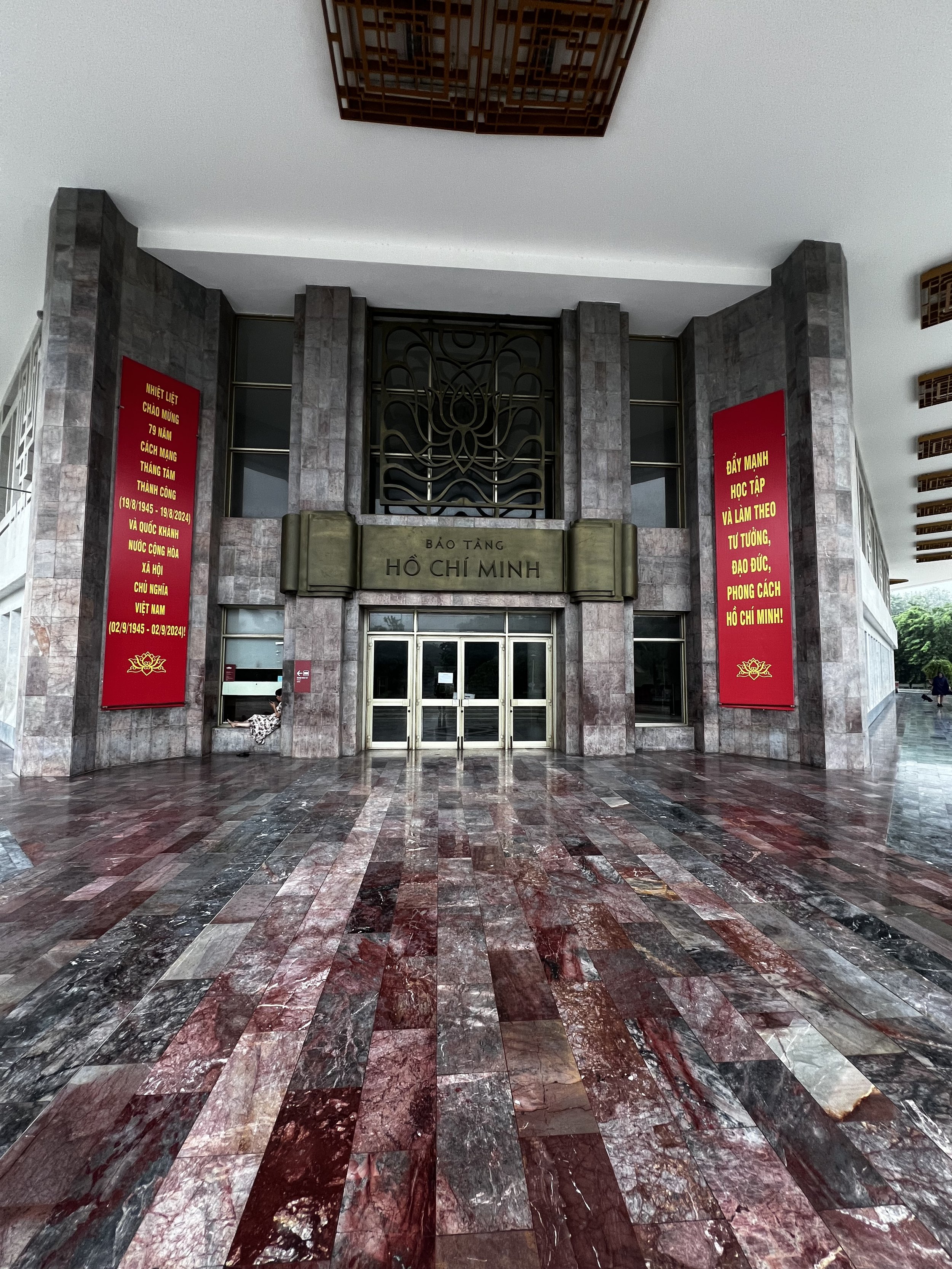 De hoofdingang van het Ho Chi Minh Museum in Vietnam met een grote metalen plaquette met de naam en rode banners met gele tekst aan weerszijden.