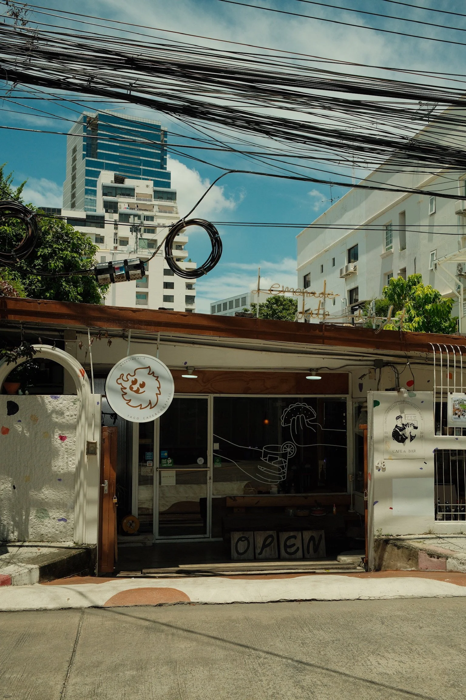 Een kleine café-bar met een open deur en decoraties op de gevel, waaronder een teken van een hand die een spraakwolk vasthoudt, een logo met een gezicht en een krullend haar, en een open teken. Boven de bar hangen elektrische kabels en er staan hoge gebouwen in de achtergrond onder een blauwe lucht met enkele wolken.