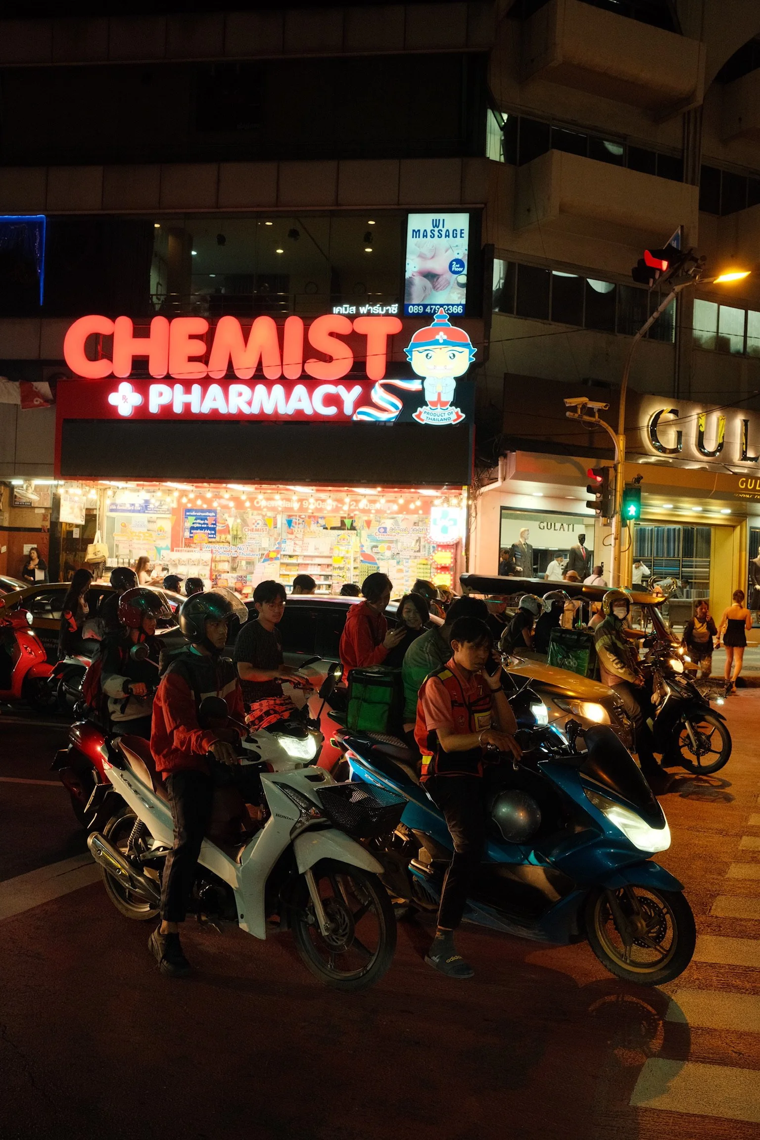 Een drukke straat met motorfietsen en scooters die wachten bij een reclamebord van een apotheek 'Chemist Pharmacy' met neonverlichting in een stedelijk gebied 's avonds.