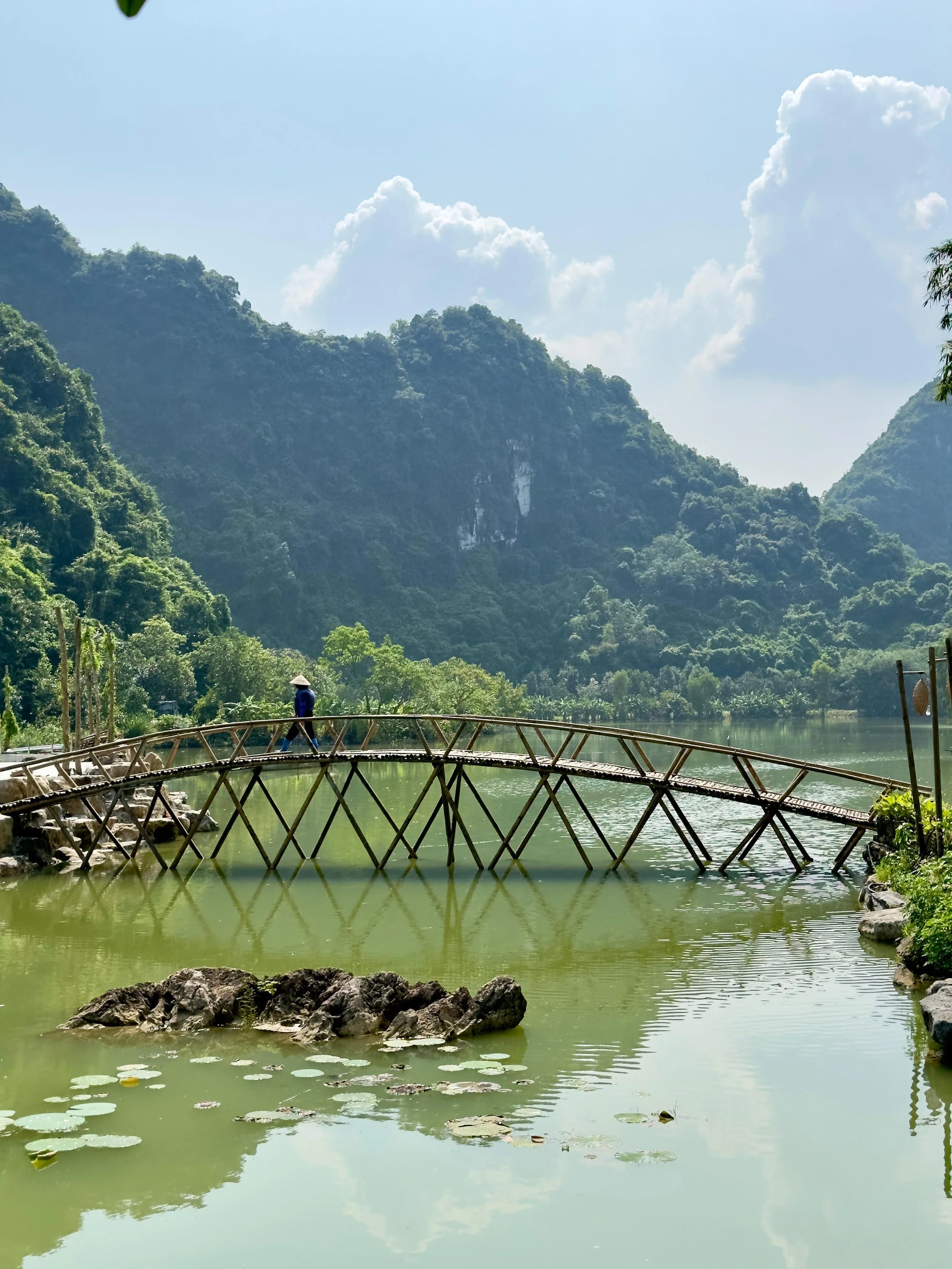Bamboebrug over een groene rivier, omringd door groene bergen en een blauwe lucht met witte wolken.