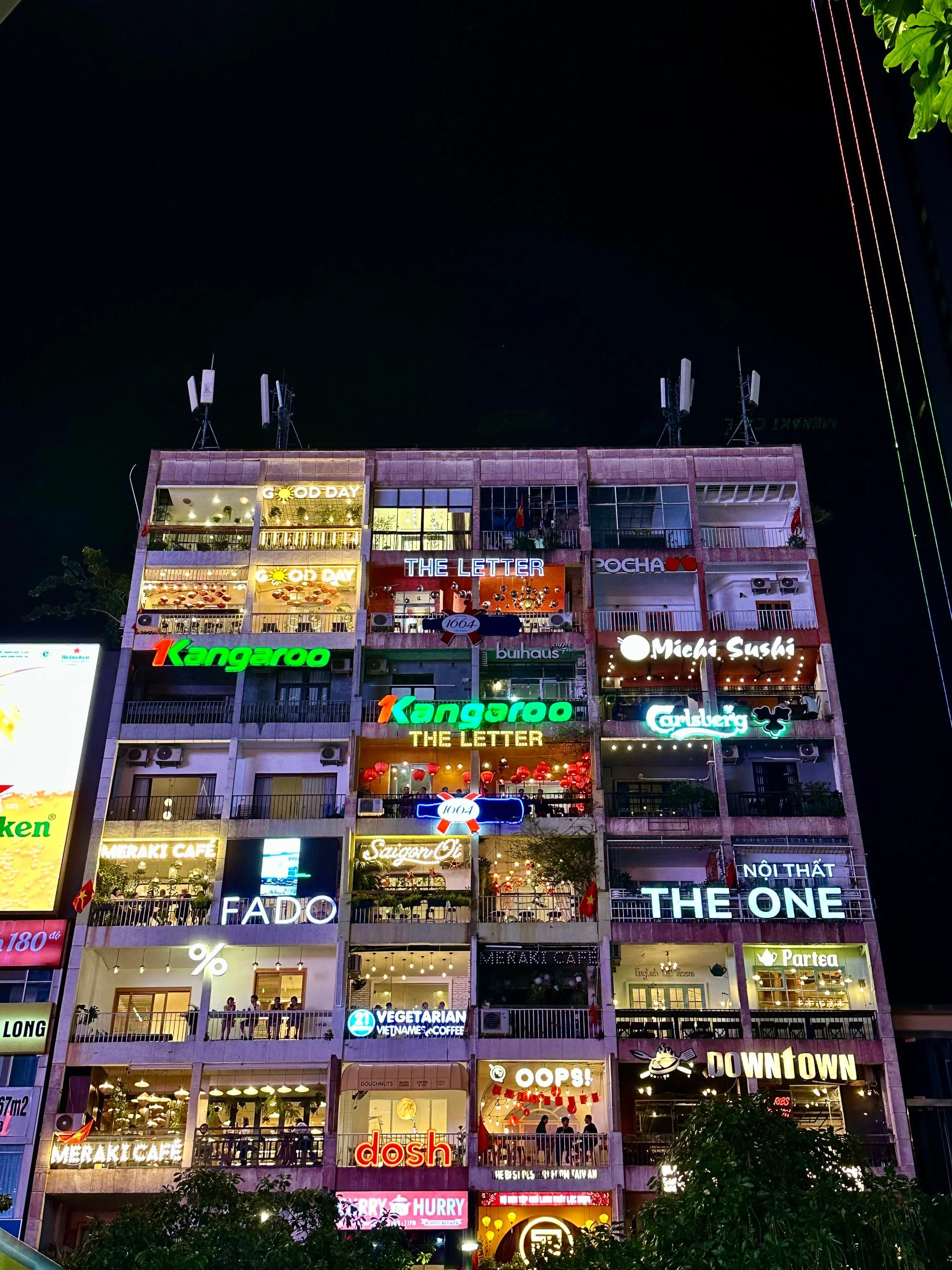 Een hooggebouw met meerdere etages, versierd met verschillende neon- en lampenkัยlen van restaurants, cafés en winkels, met een donkere avondhemel op de achtergrond.
