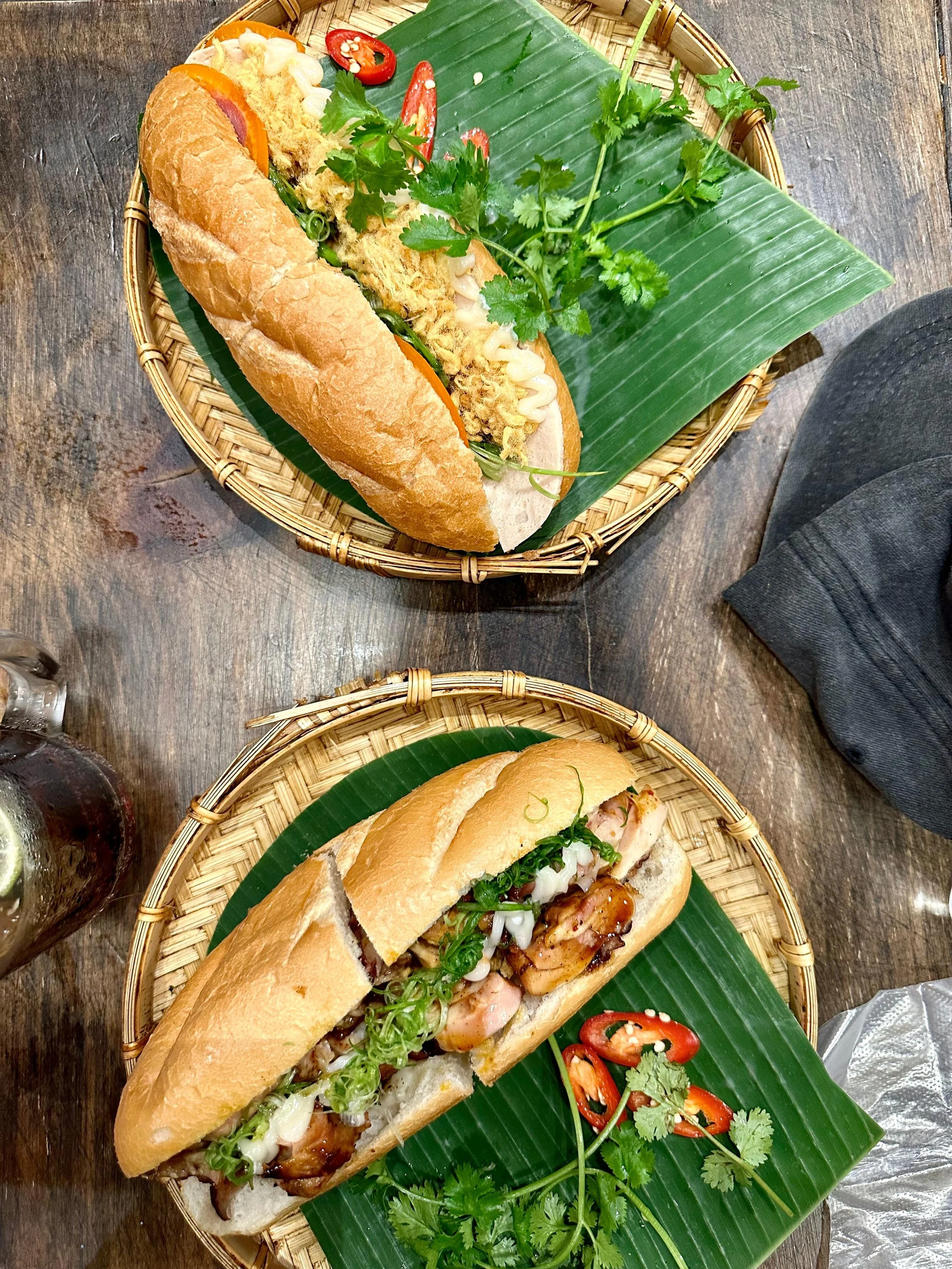 Twee broodjes Banh Mi op een houten tafel, elk op een dienblad met een groene blad en wat chilipepers en koriander als garnering.