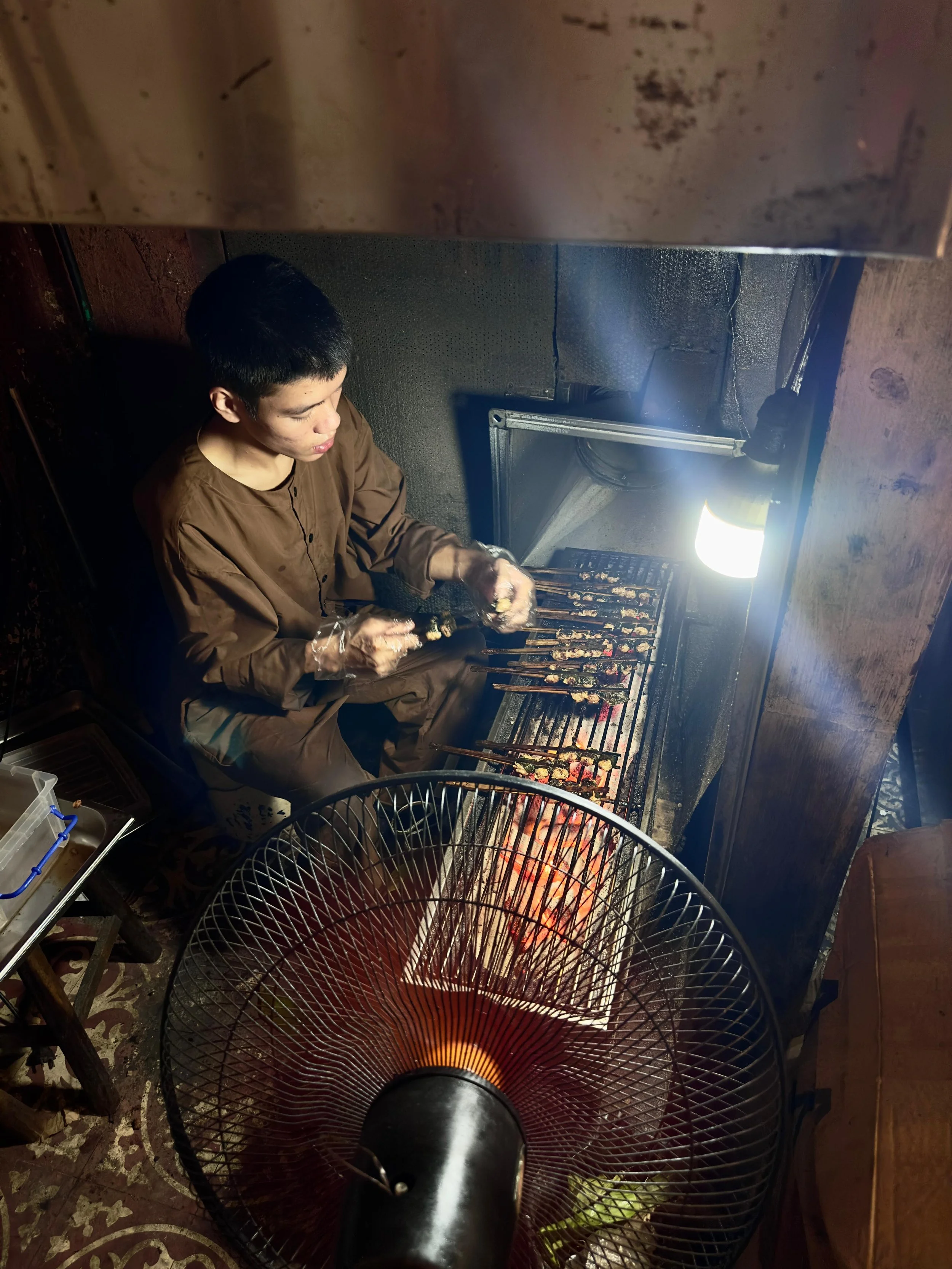 Man die saté's grilt op een barbecue, met een ventilator in de voorgrond.