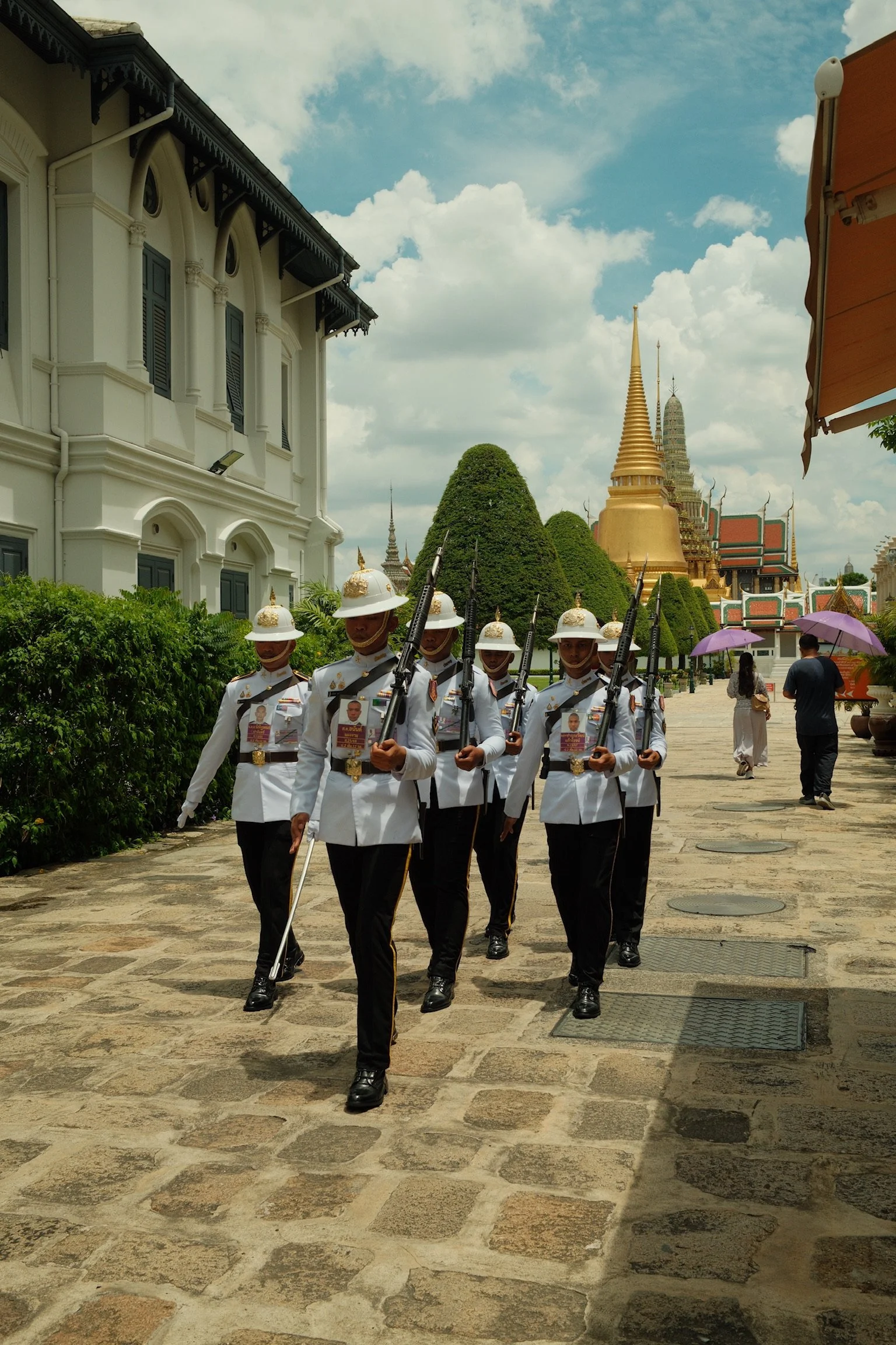 Een lid van het koninklijke militaire personeel marcherend in een ceremonie rondom een tempel, met een opvallende gouden stupa op de achtergrond, onder een bewolkte hemel in Thailand.