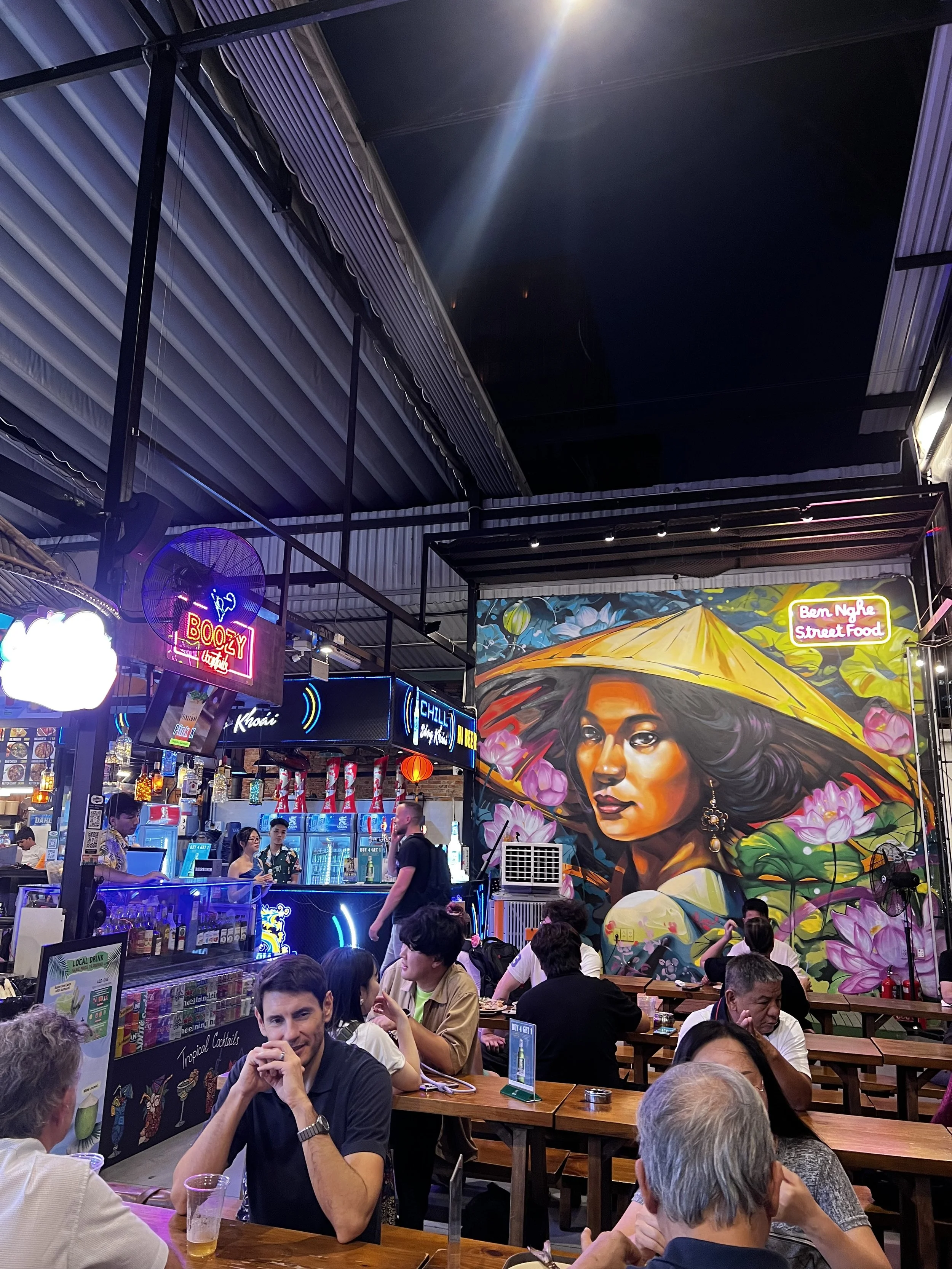 Interieur van een restaurant of bar met klanten aan tafels, een kleurrijke muurschildering van een vrouw met een grote gele hoed en bloemen op de achtergrond, neonborden, bar met drankjes en televisies.