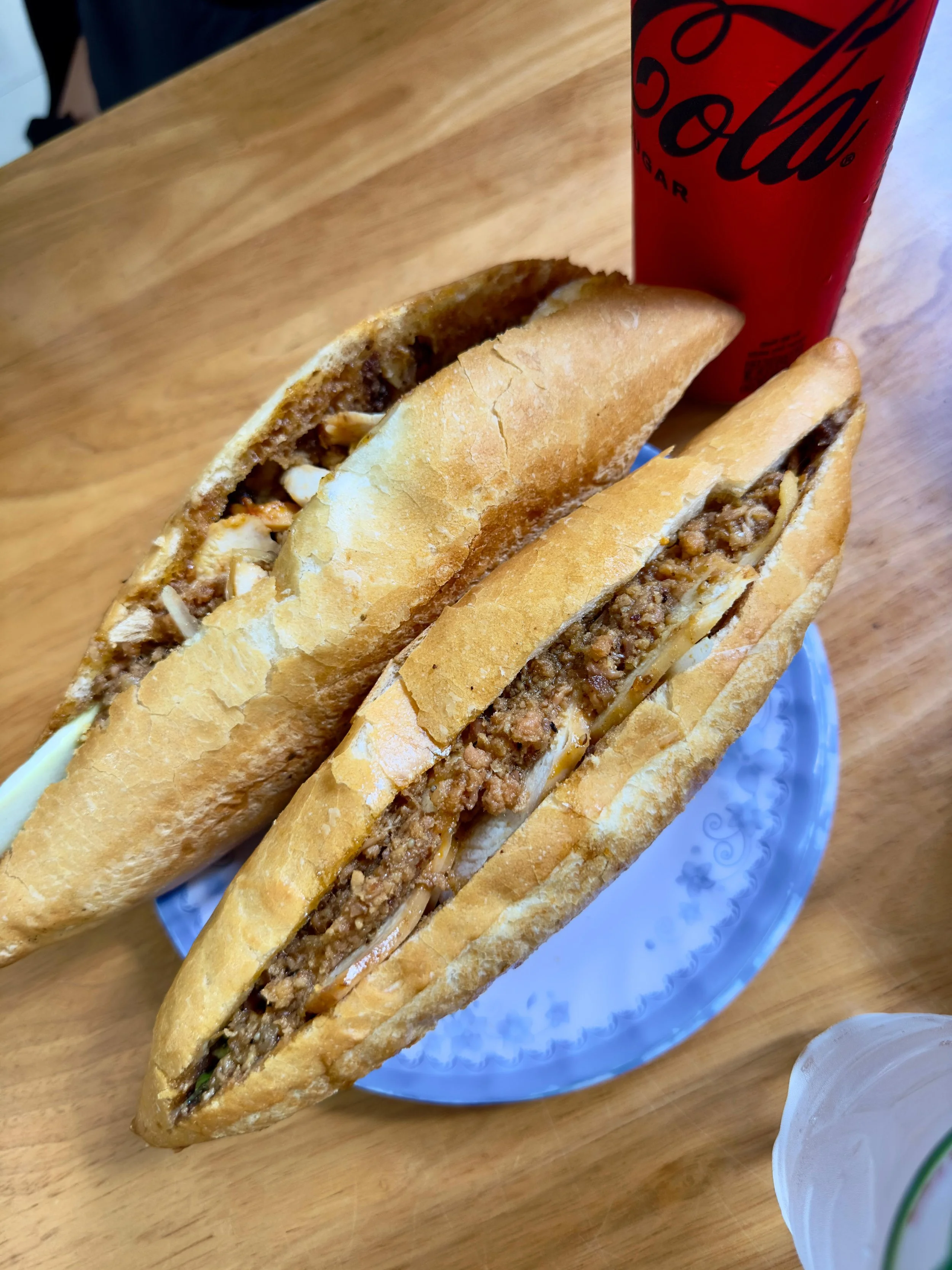 Twee Bành Mì broodjes met gekruid gehakt en kaas, op een blauwe bord, met een fles Coca-Cola en een glas op een houten tafel.
