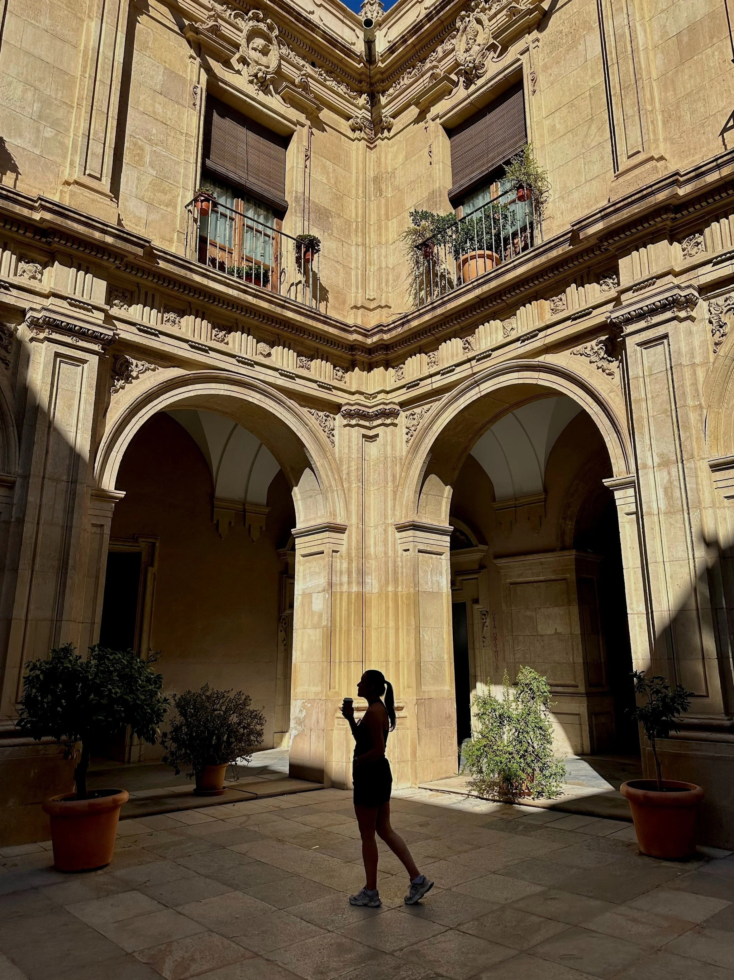 Silhouet van een vrouw met een drinkbeker in een binnenplaats met historische gevel en planten