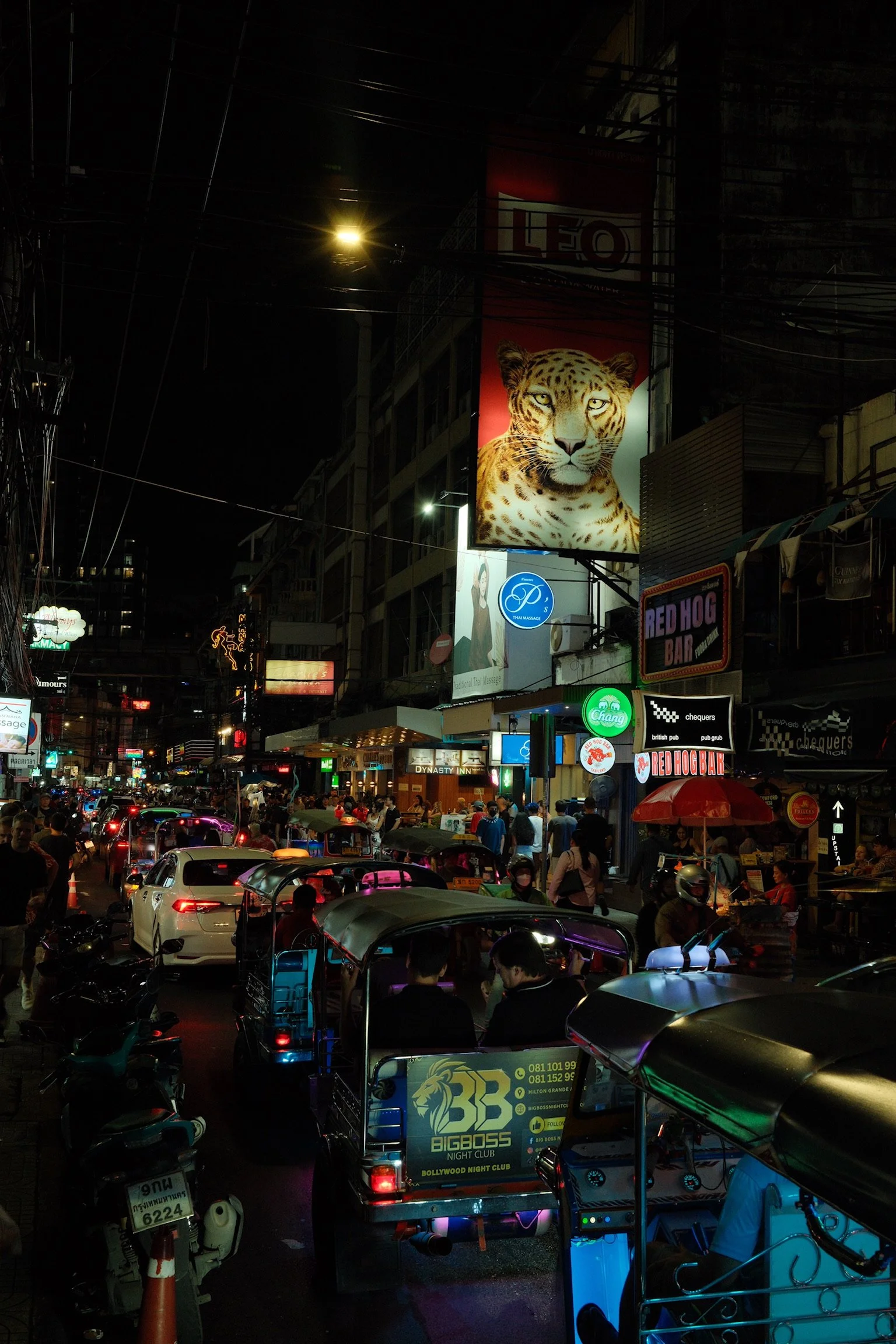 Een drukke straat 's avonds met veel auto's, motoren en mensen. Verlichte winkels en neonborden, met een grote advertentiebanner van een luipaard en andere reclames.