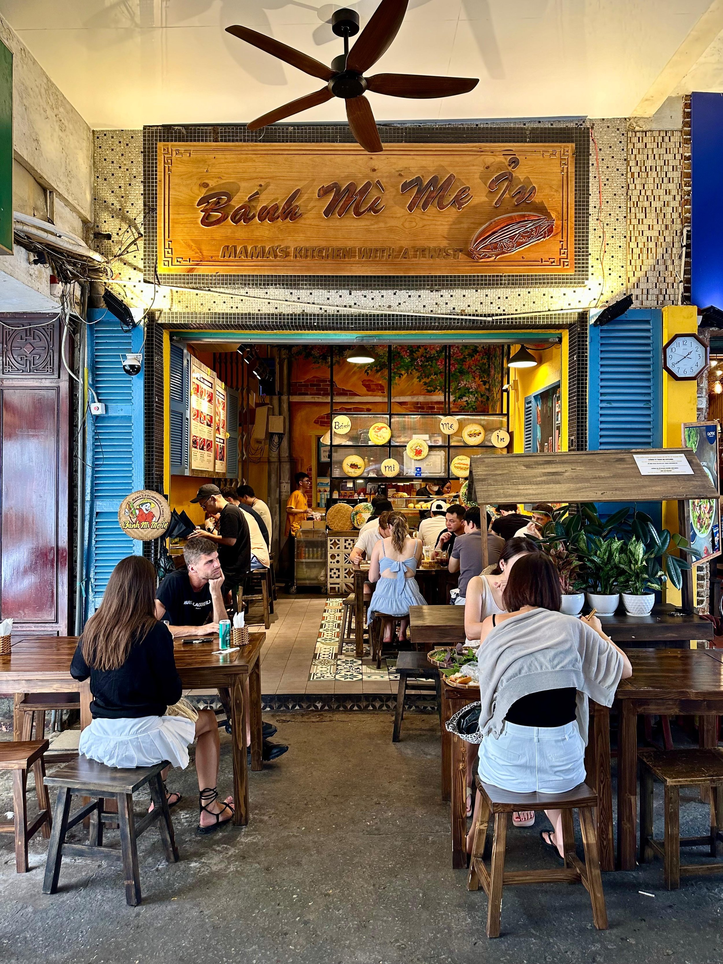 Gezellig Vietnamees restaurant met mensen die genieten van hun maaltijd. Het interieur heeft warme kleuren en decoraties, met grote pannen en een menukaart aan de muur.