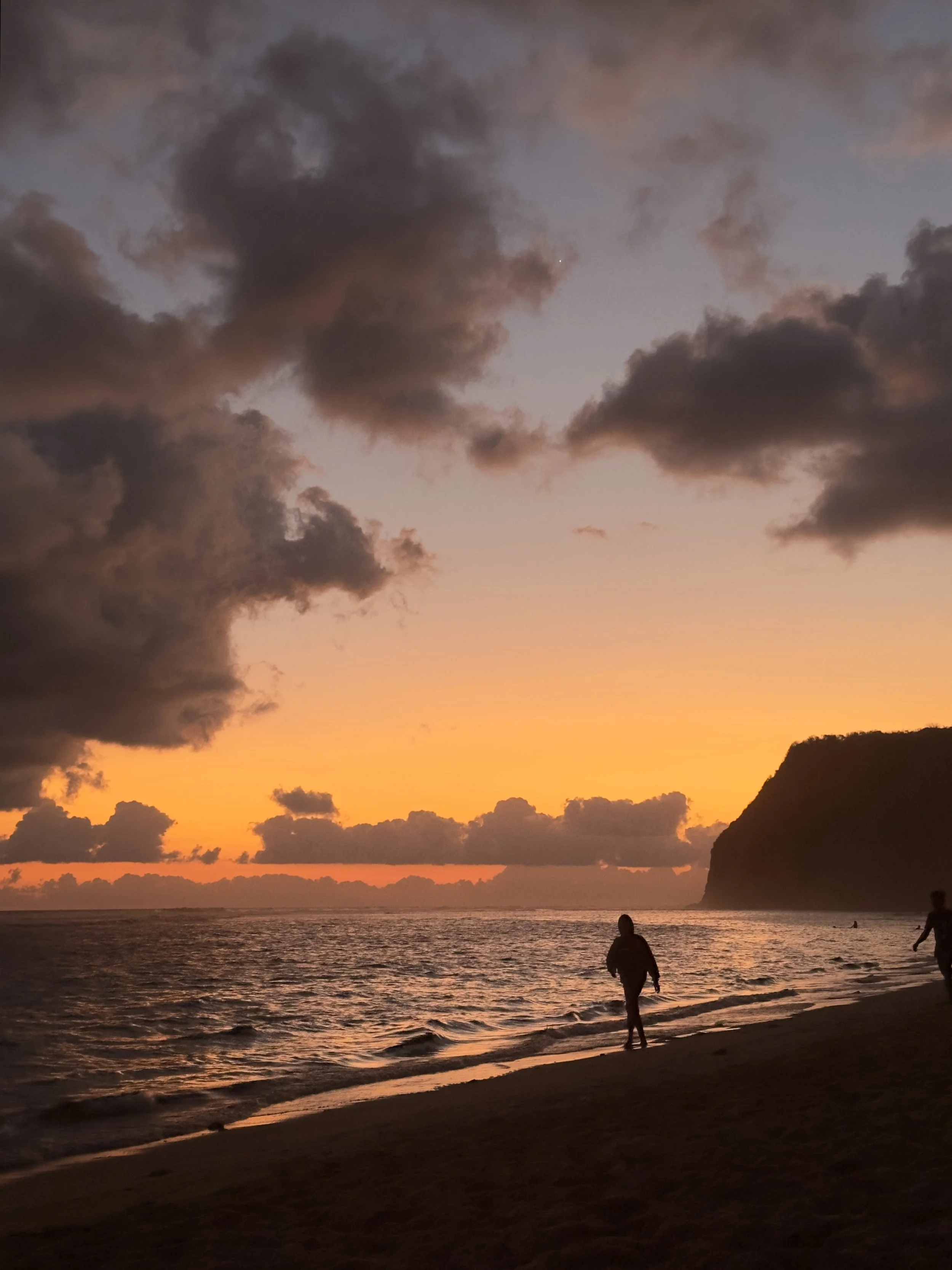 Silhouetten van mensen die op het strand lopen bij zonsondergang, met een rotsachtige klif op de achtergrond en een bewolkte hemel.