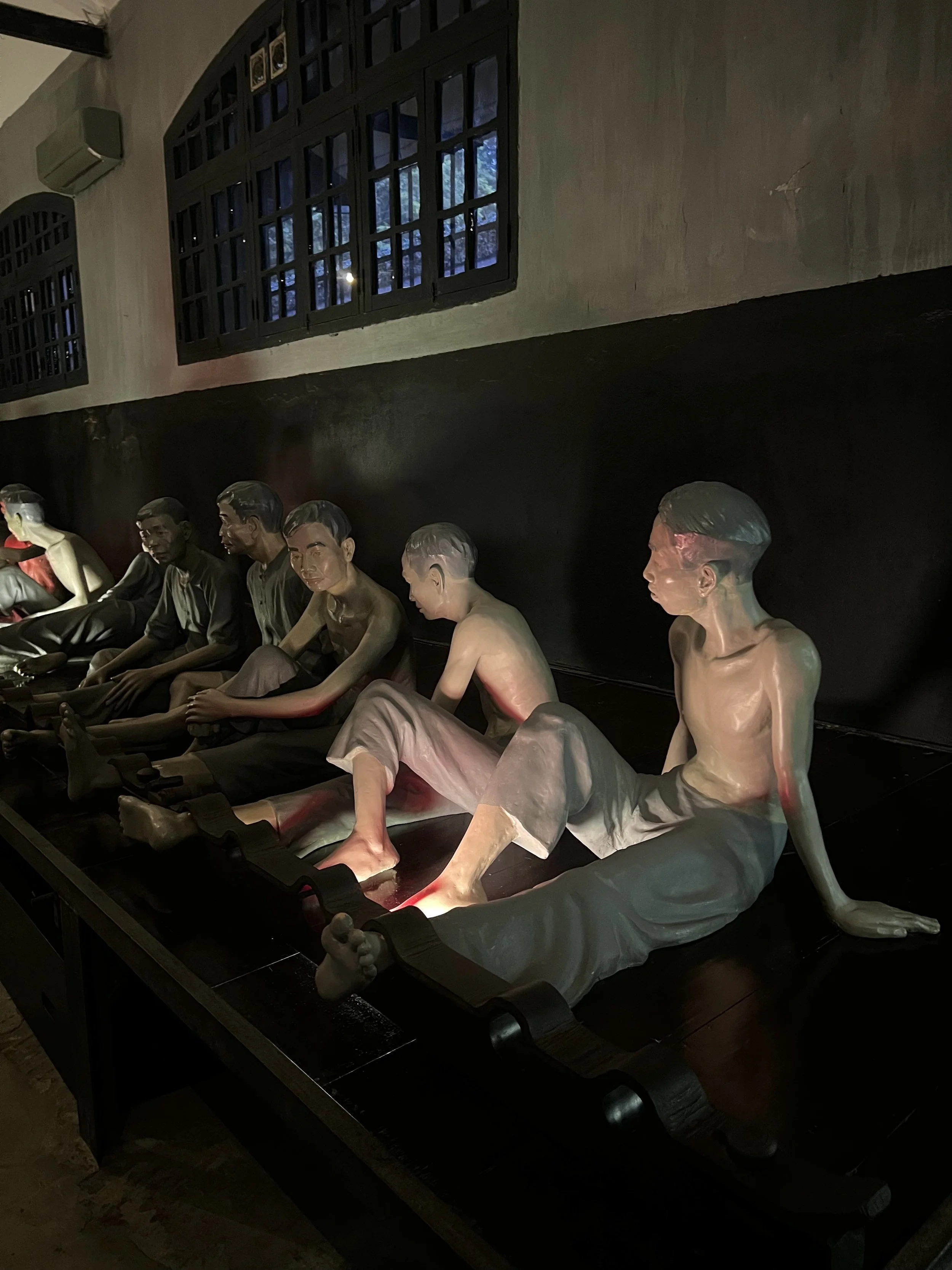 Beelden van levenloze replica's van gestileerde mensen, waarschijnlijk geteeld voor educatieve of herdenkingsdoeleinden, in een donkere ruimte met ramen en muren.