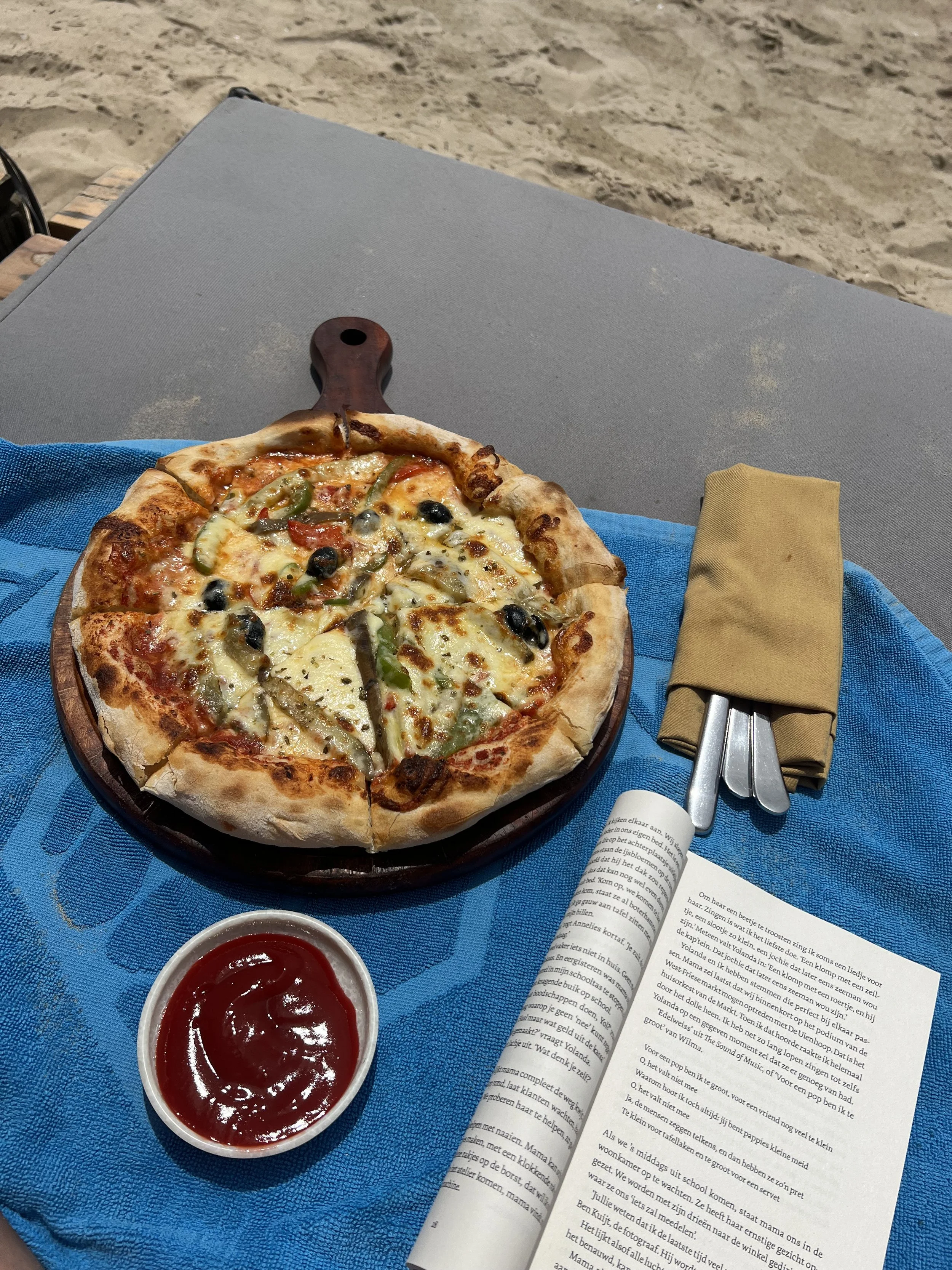Een pizza op een houten bord, een kom met ketchup, bestek ingepakt in een okerkleurige servet, en een open boek op een blauwe tafelkleed, buiten met zand op de achtergrond.