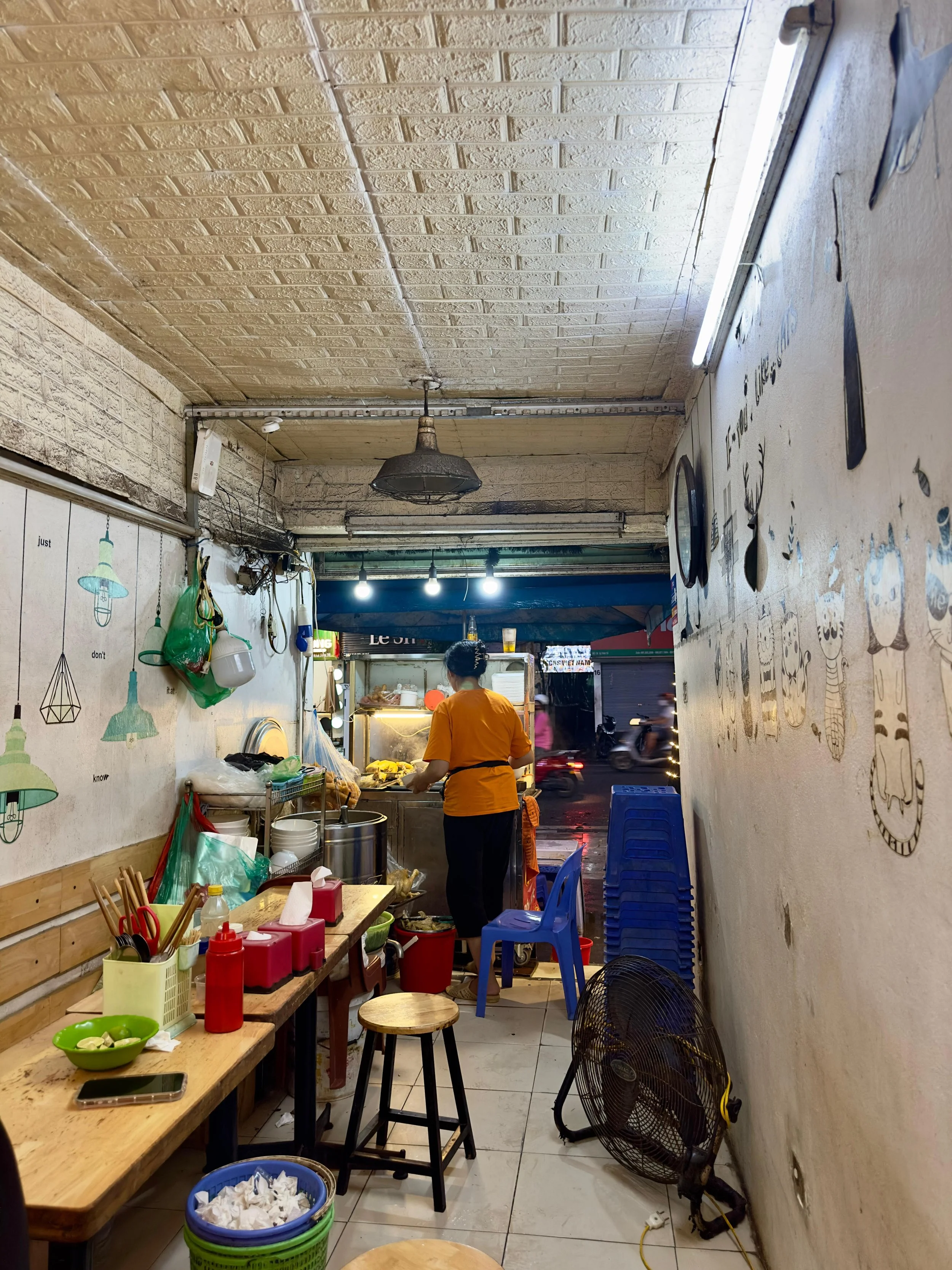 Binnenkant van een klein restaurant met een persoon die aan een kookstation staat. Taferelen met stoelen, tafels, keukenbenodigdheden, en een ventilator.