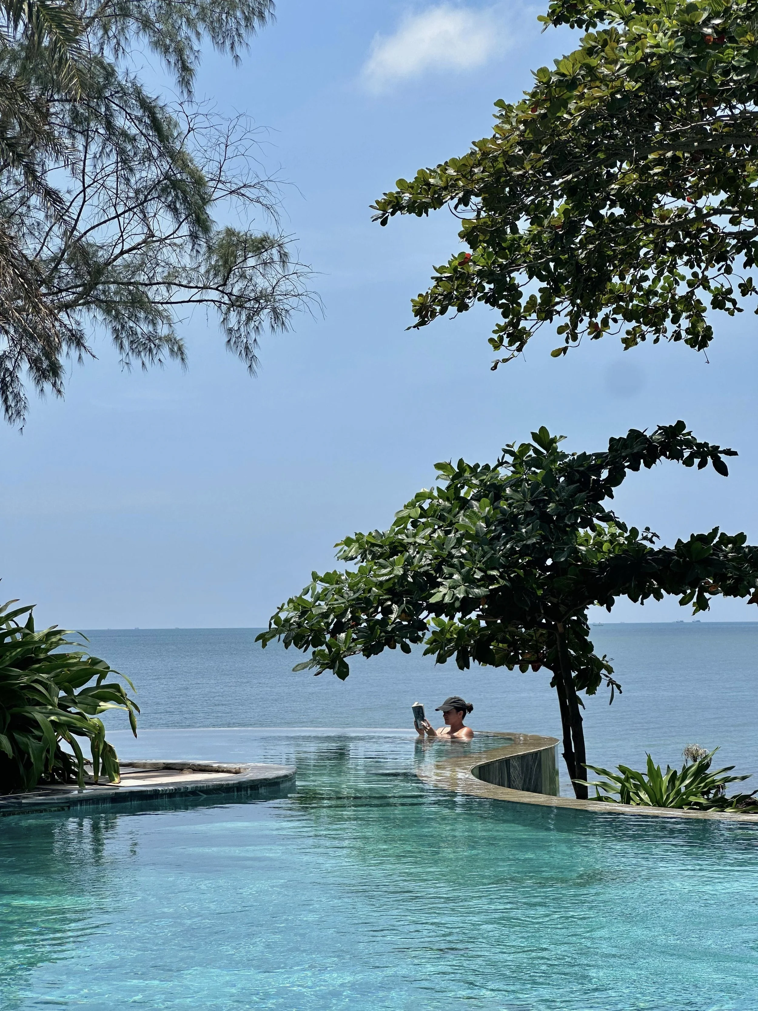 Persoon met pet die in zwembad een boek leest, omgeven door groene planten, met zee op achtergrond en blauwe hemel In Phu Quoc.