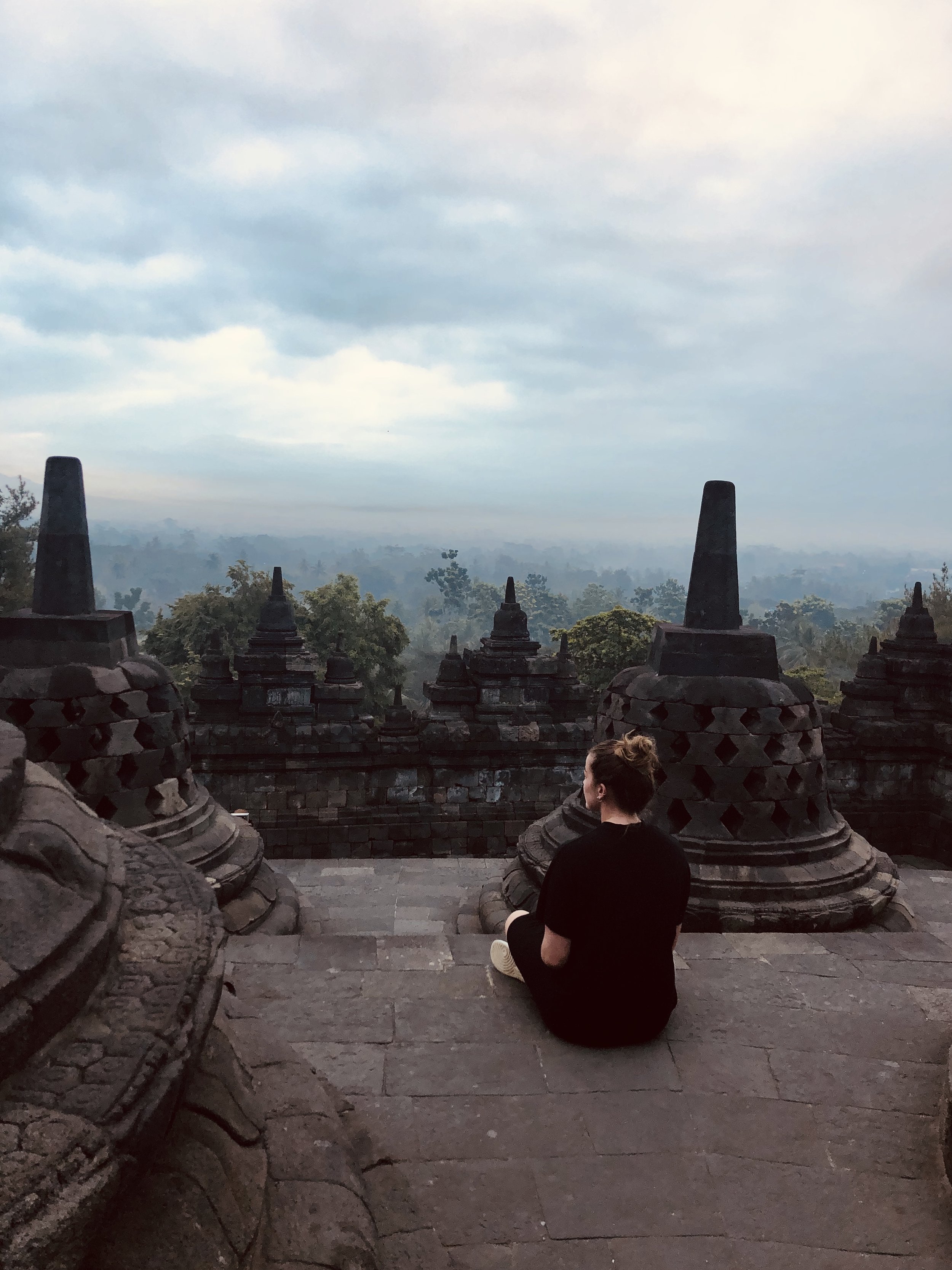 Een persoon in een zwart shirt zit op trappen tussen oude rotsstructuren, uitkijkend over een mistige groene vallei met een bewolkte hemel.