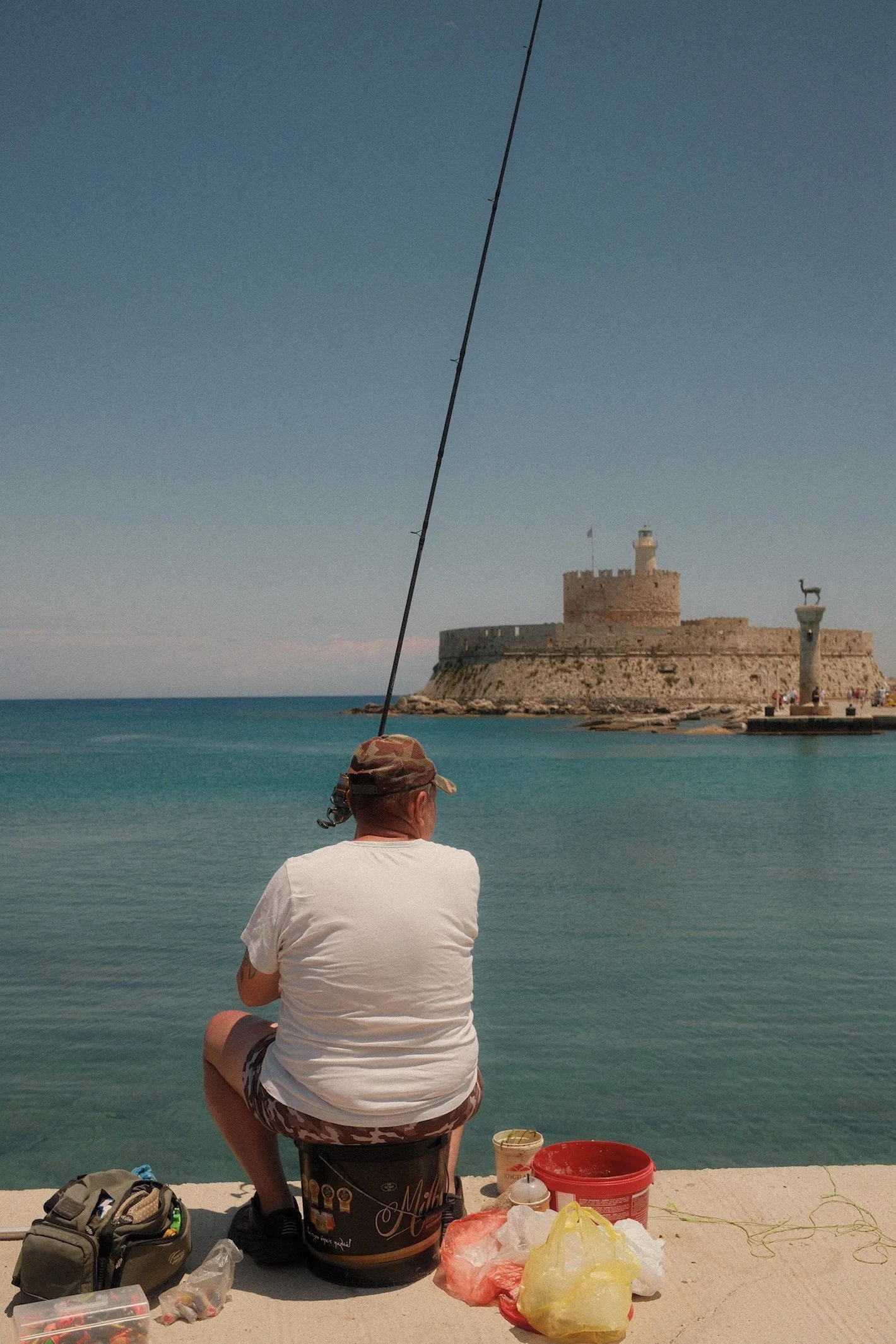 Man vissend aan de rand van een haven met een kasteel en vuurtoren op een rots in de achtergrond.