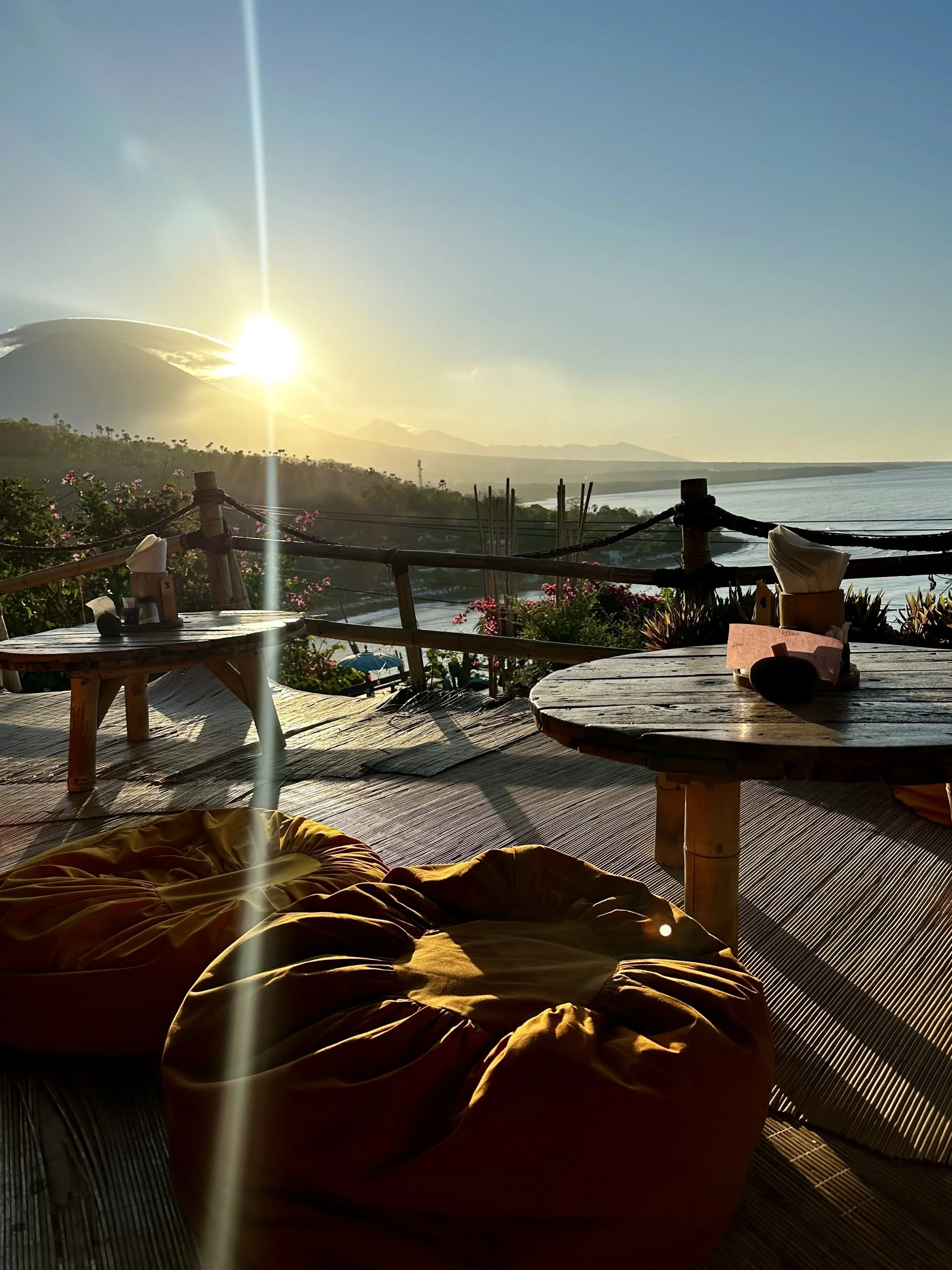 Op een terras met kussens en lage houten tafels, uitzicht op de zee en bergen met de zon ondergaat achter de horizon.