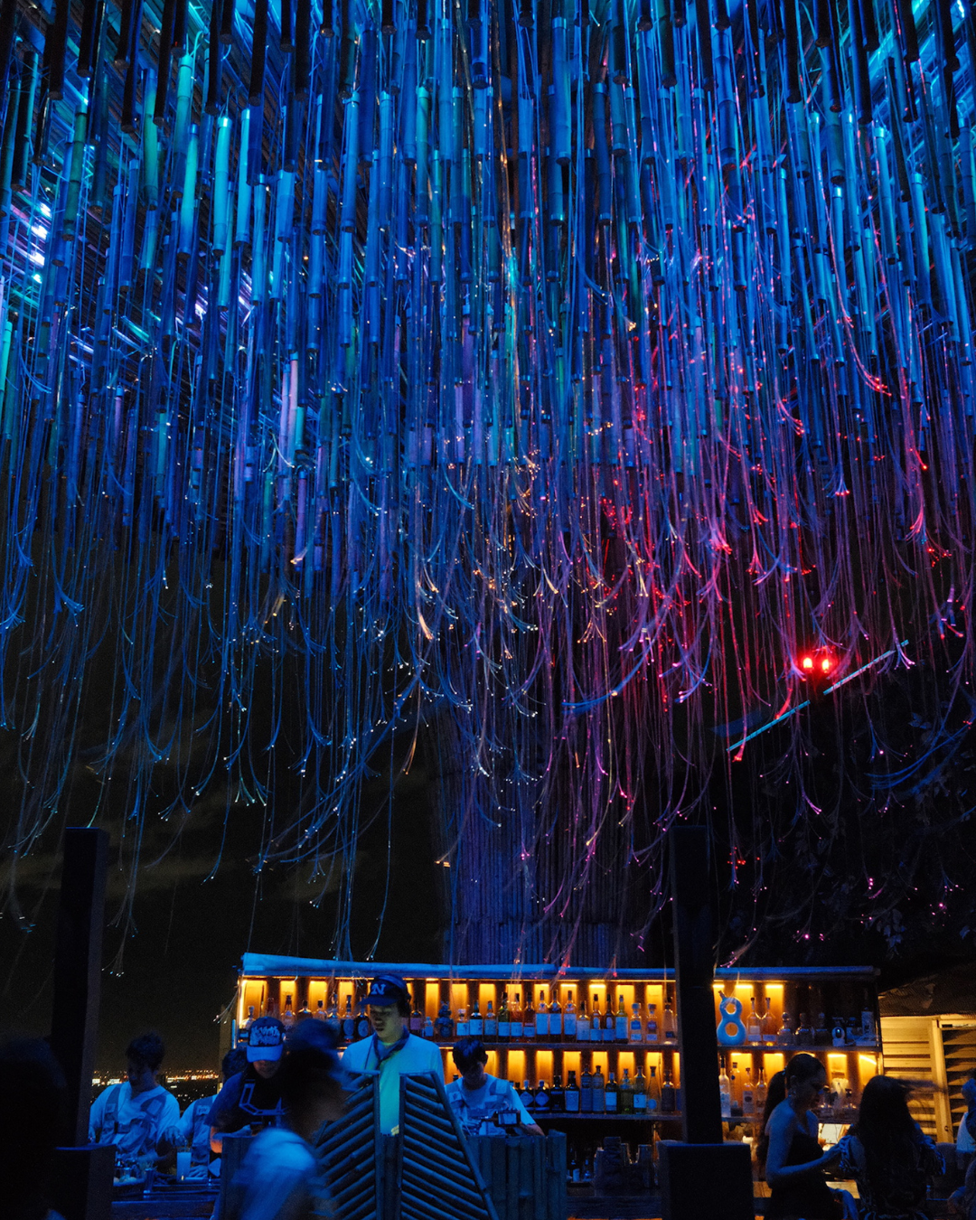 Een groep mensen die genieten van een nachtelijke gebeurtenis onder een kleurrijk kunstinstallatie van geleidende draden en LED-verlichting, met een bar en snacks op de voorgrond.