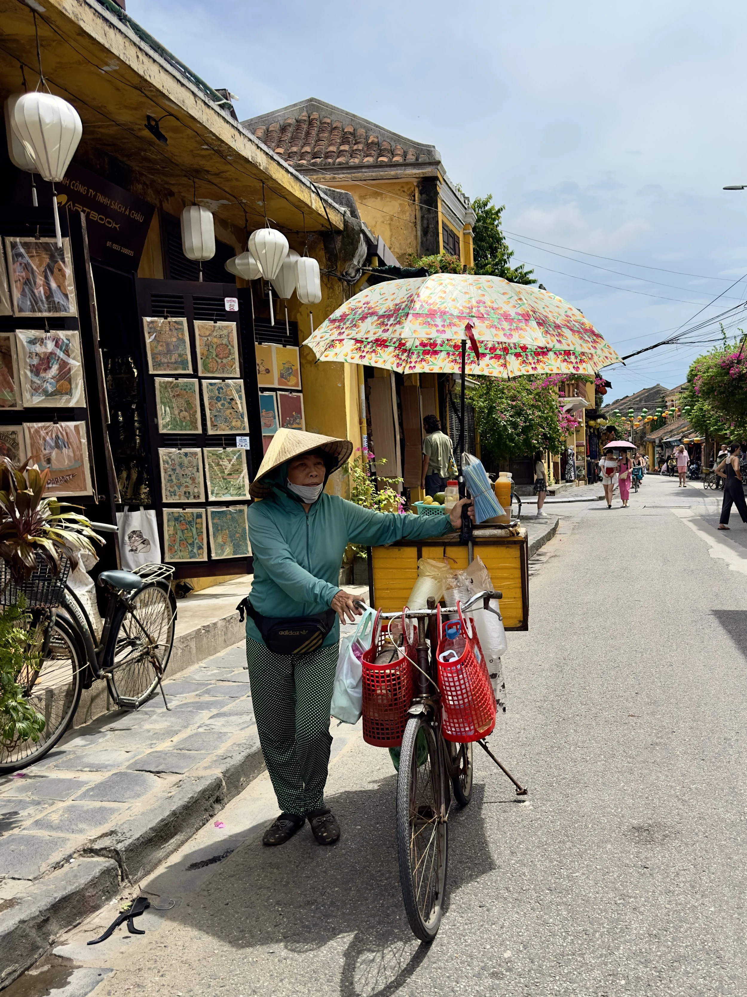 Vietnamese vrouw met een hoed verkoopt snacks vanaf haar fiets met een kleurrijke paraplu in een drukke straat in een stad in Hoi An.