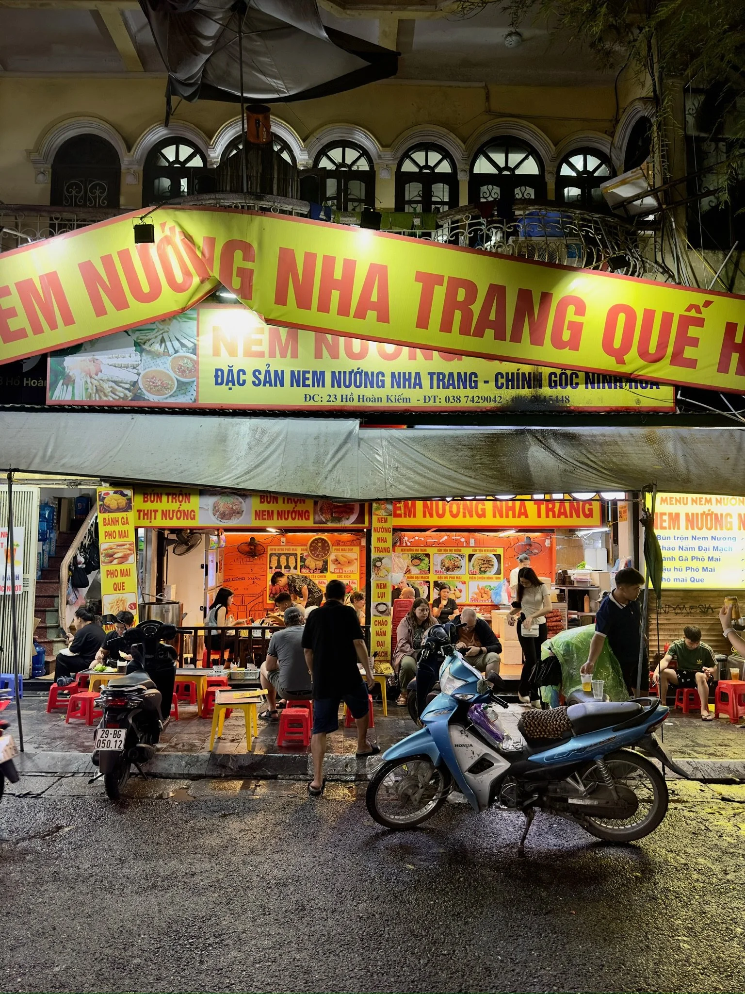 Een Vietnamese straatkraam die nem nướng serveert met mensen die daar eten en passanten op de natte straat.