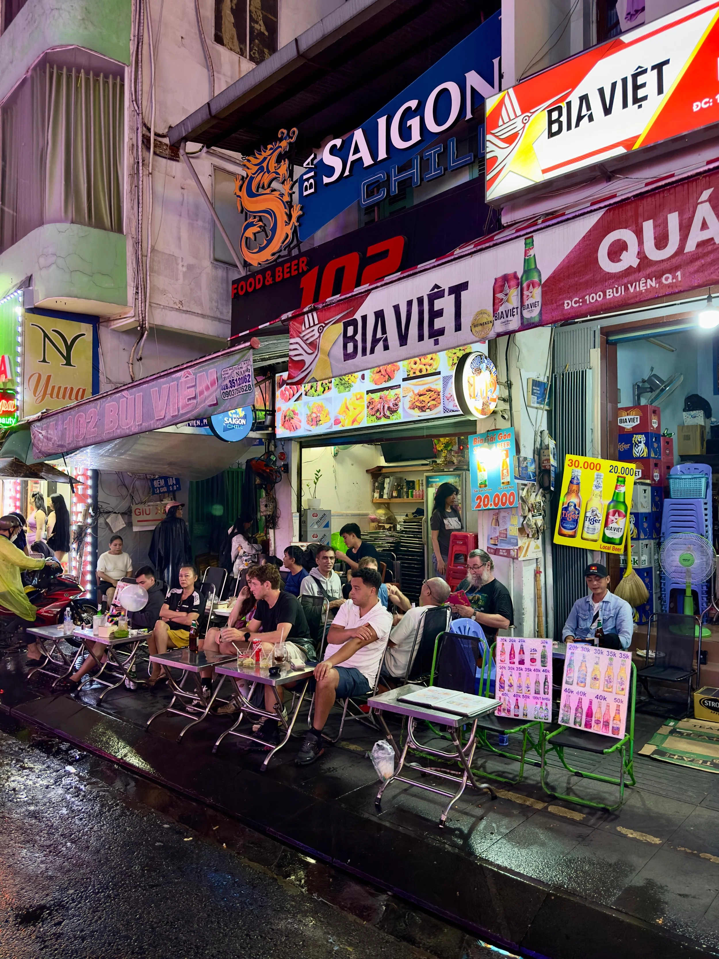 Sfeervolle straatkant met een restaurant waar mensen zitten te genieten van een avondmaaltijd op een regenachtige avond, met neonverlichting en kleurrijke signboards.