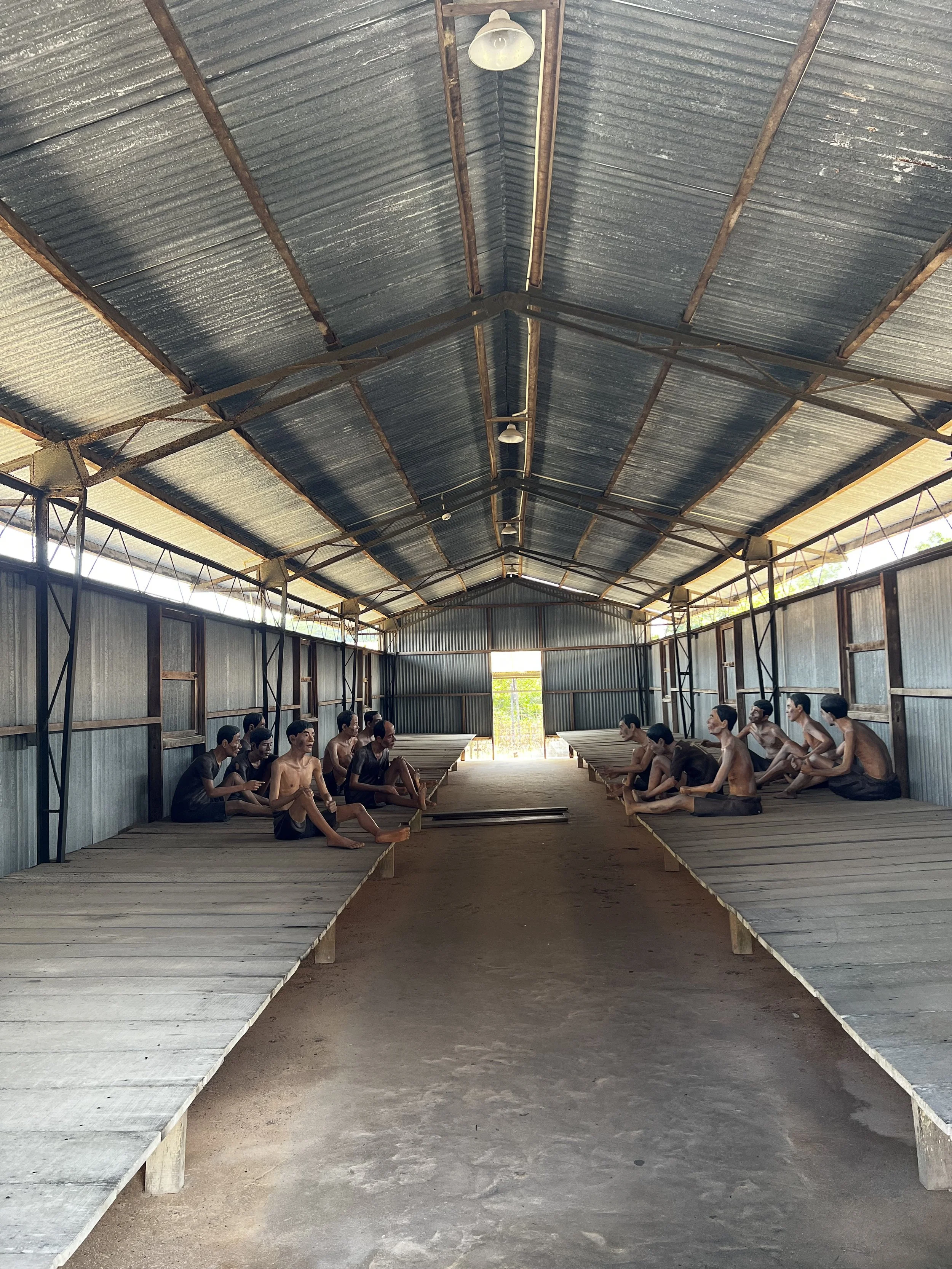 Binnenkant van een eenvoudige houten structuur met hoge metalen plafond en zitbanken aan beide zijden, met bewerkte beelden van mensen die op de banken zitten.