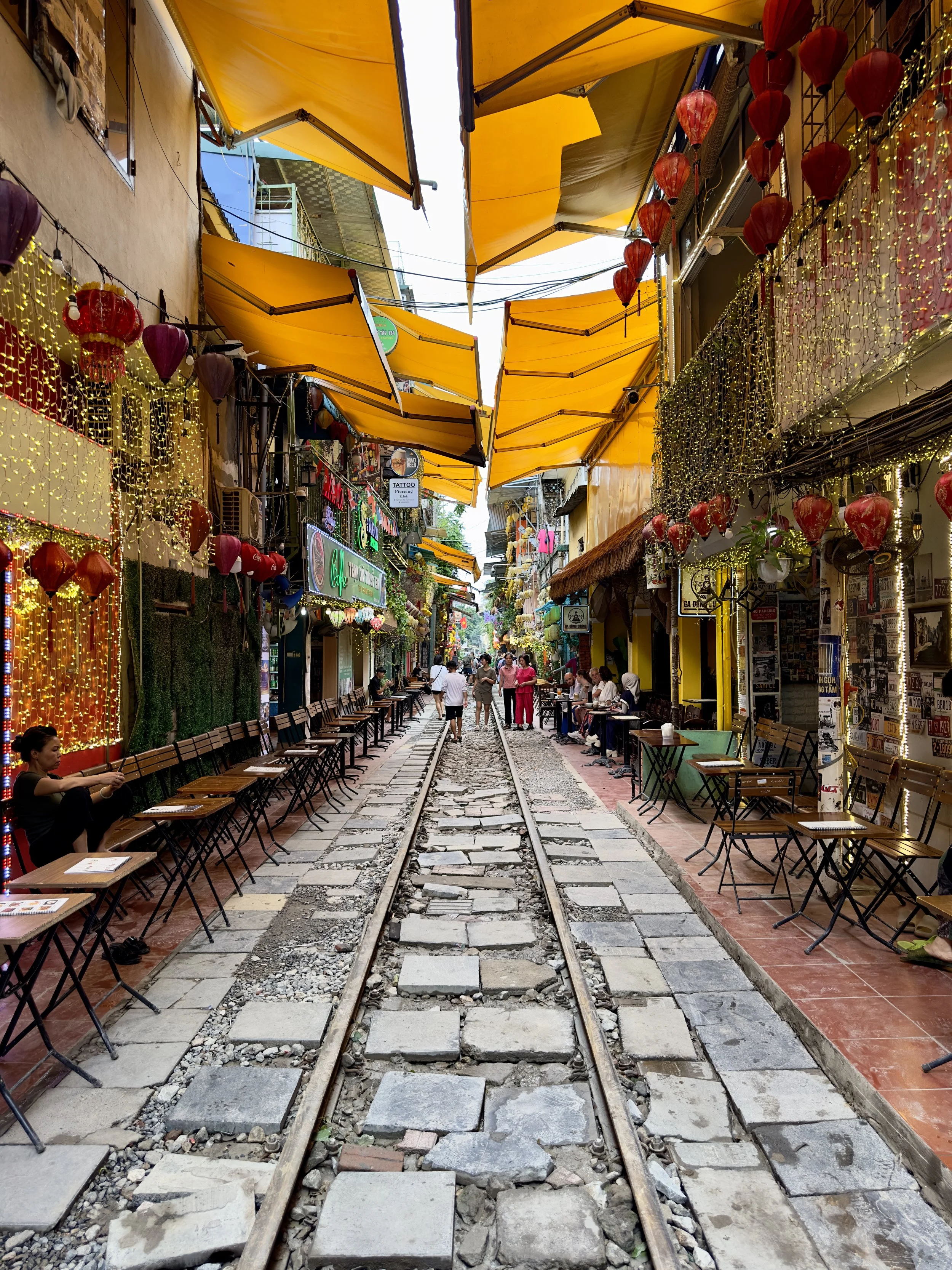 Een kleurrijk straatje met een spoorlijn in het midden, omgeven door cafés en winkels, versierd met rode en paarse lantaarns en gele markiezen.