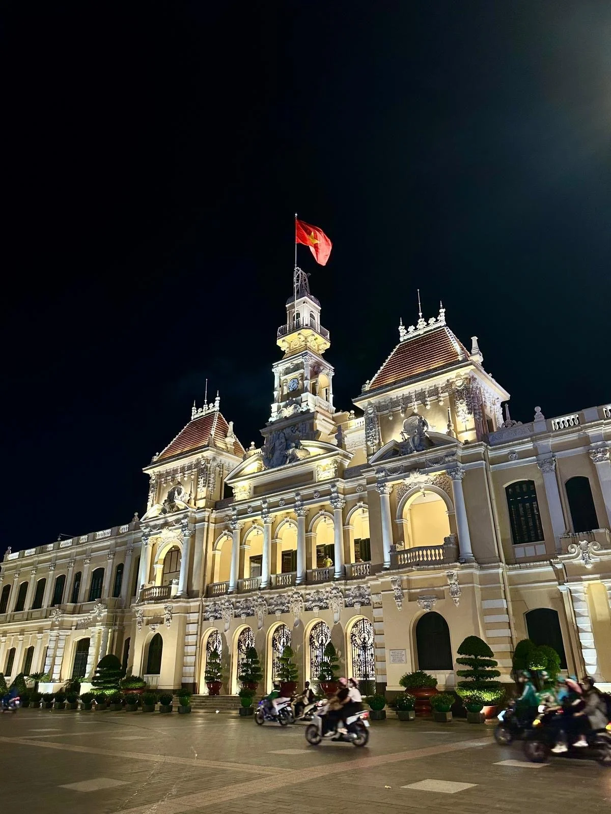 Een verlicht barok paleisachtig gebouw met een vlag die wapperd op een toren, tegen een nachtelijke hemel, met motorfietsen die voorbijrijden op de straat voor het gebouw.