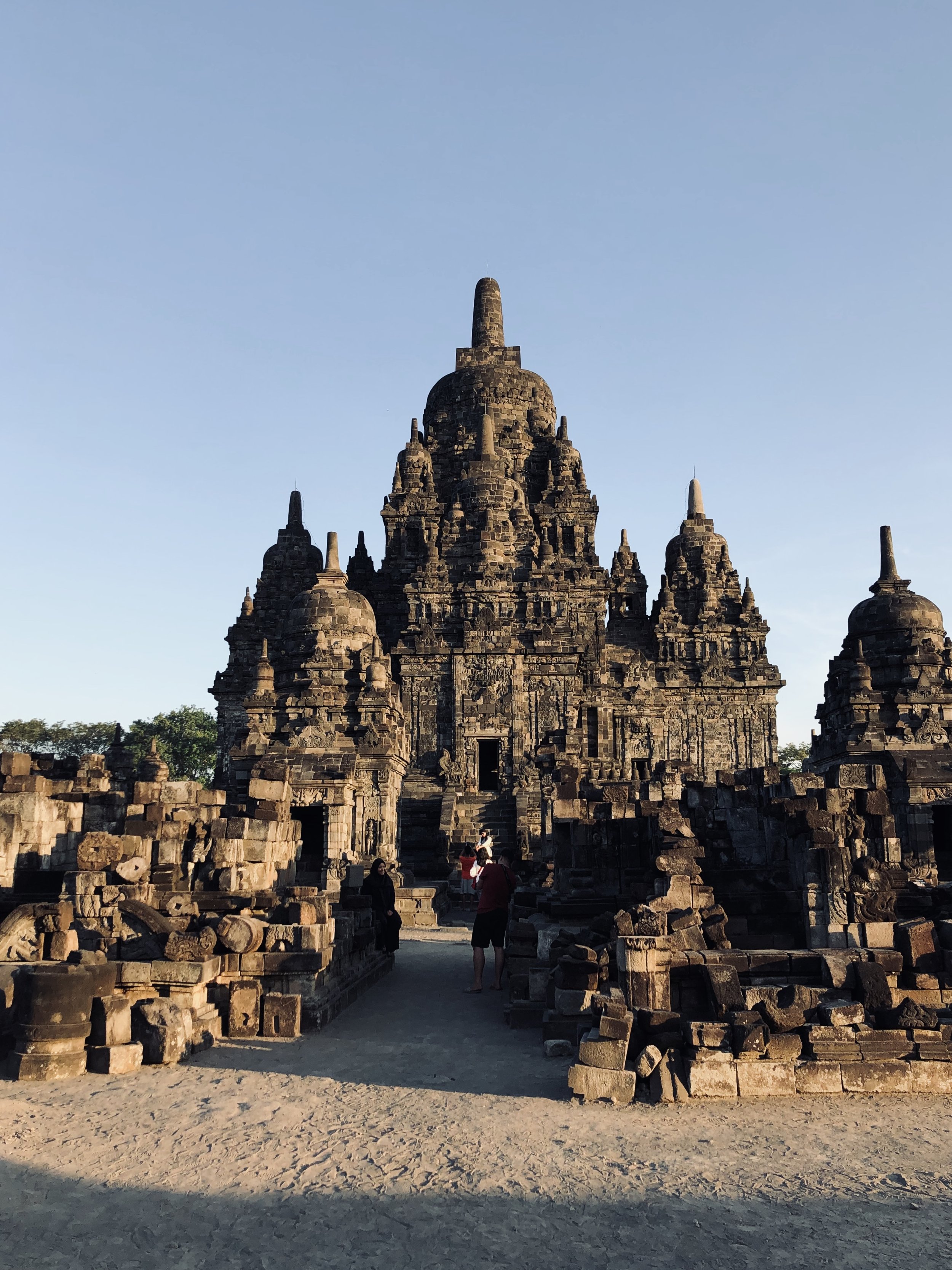Een oude stenen tempel met meerdere torens en beeldhouwwerken, omgeven door een zandpad en een paar mensen die eromheen lopen