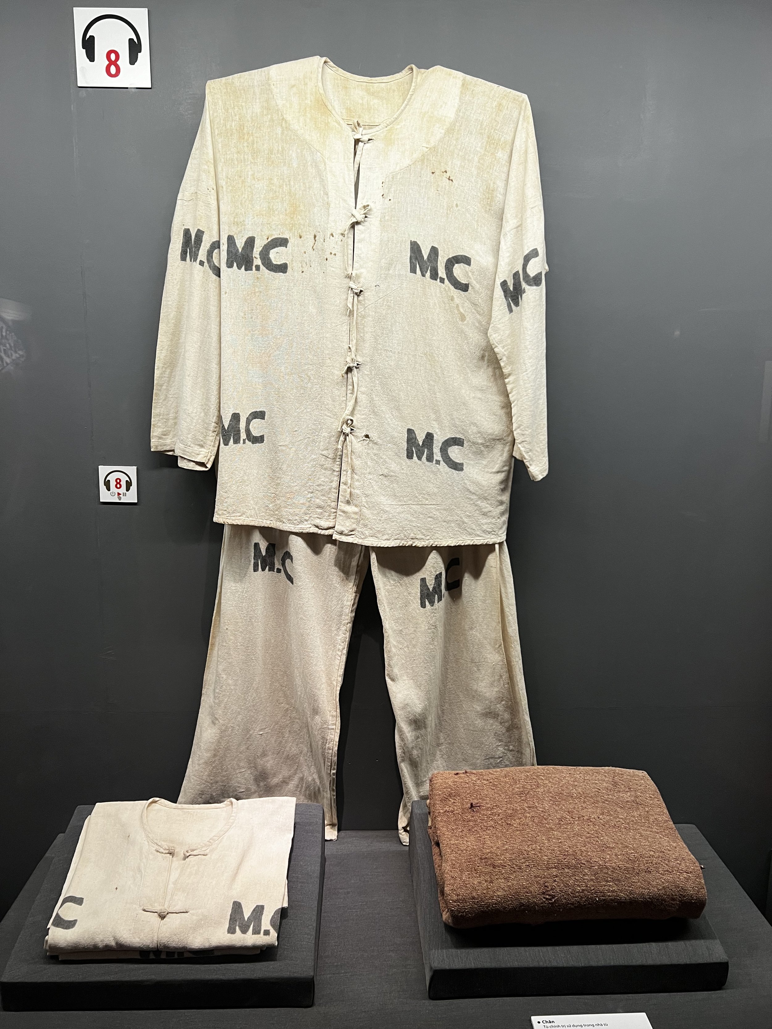 Een beige huishoudoutfit bestaande uit een jas en broek, met zwarte letters 'M.C' en 'N.C.M.C' erop. Daarnaast ligt een witte gestikte jas en een bruine lap stof op aparte planken.
