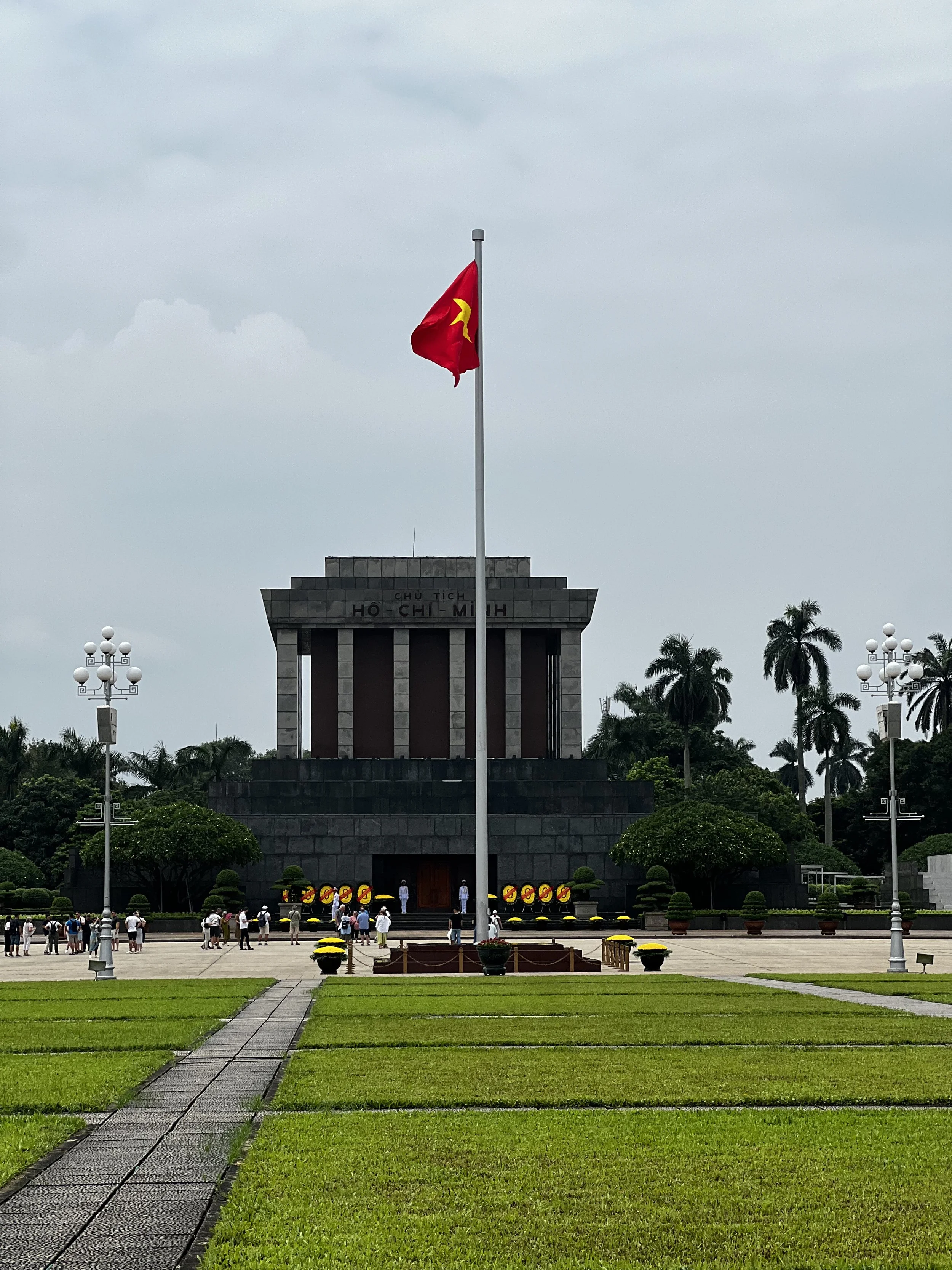 Het beeld toont het mausoleum van Ho Chi Minh met de vlag van Vietnam die wappert op de voorgrond, omringd door bomen, palmbomen en een grasveld met paden.