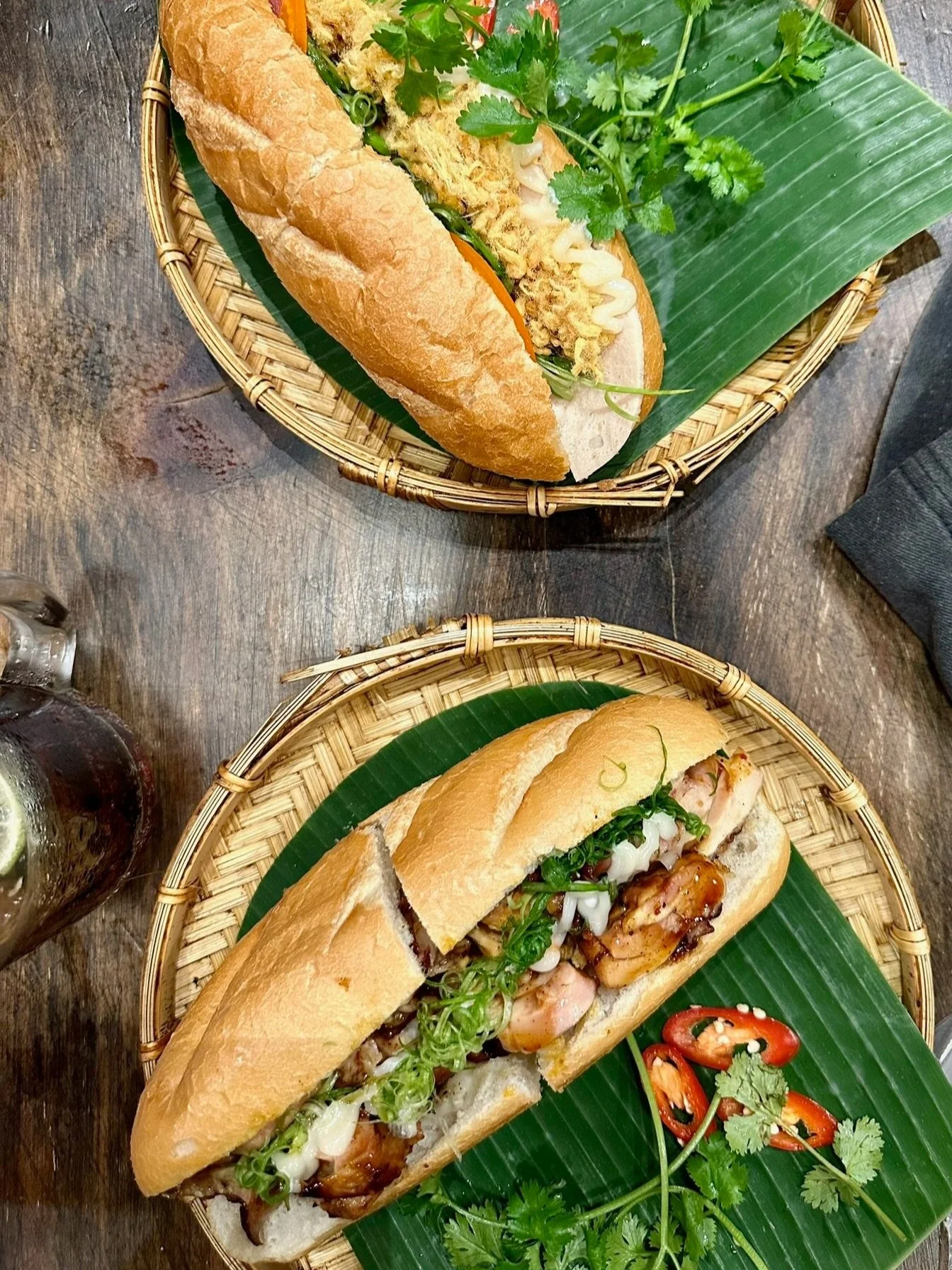 Twee sandwiches in bamboe mandjes, één met gegrilde kip, groene uien, chilipepers en koriander, op een groene bananenblad. Beide worden geserveerd op een houten tafel.