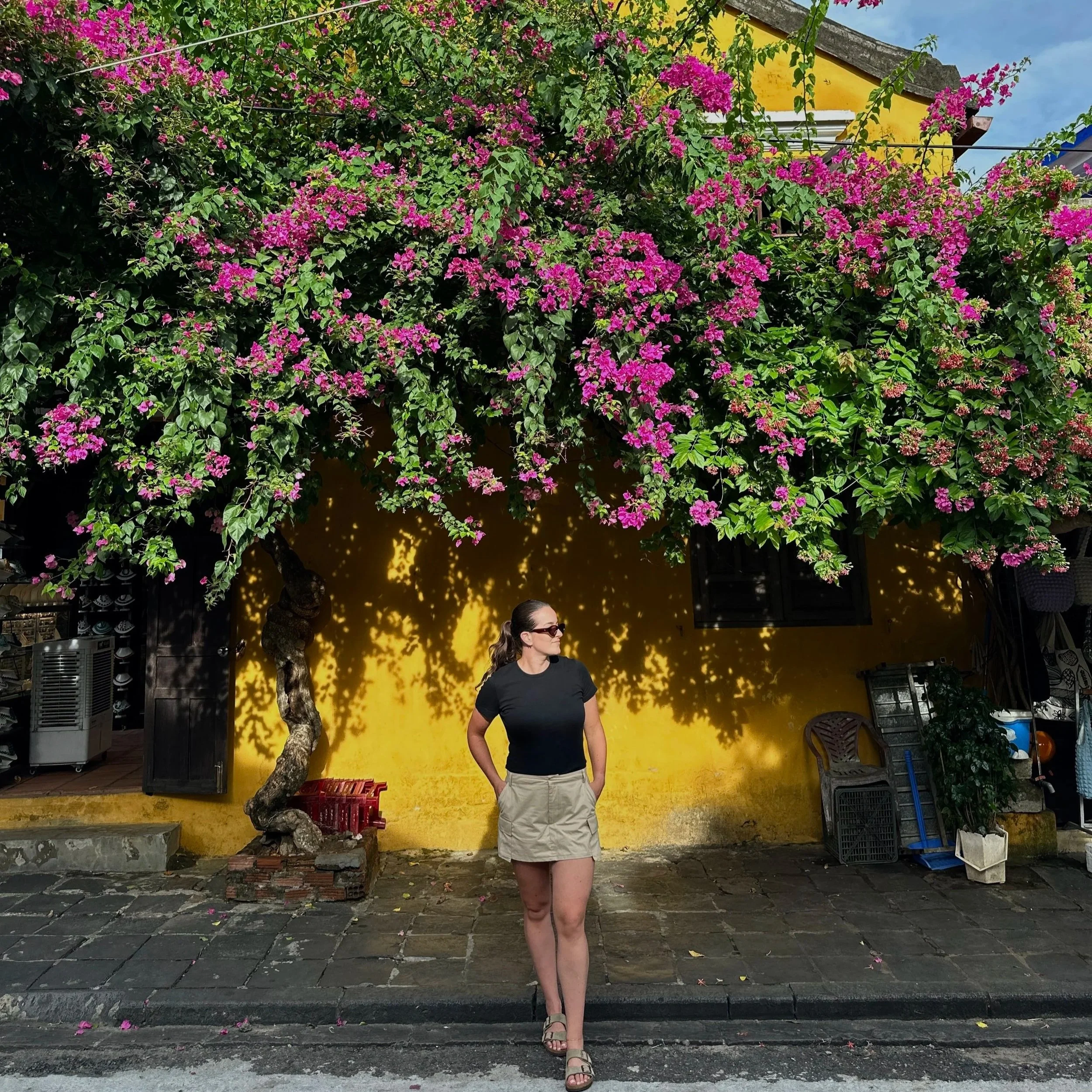 Vrouw met zonnebril voor een gele muur onder een bloeiende boom met roze bloemen in een straatje in Hoi An.