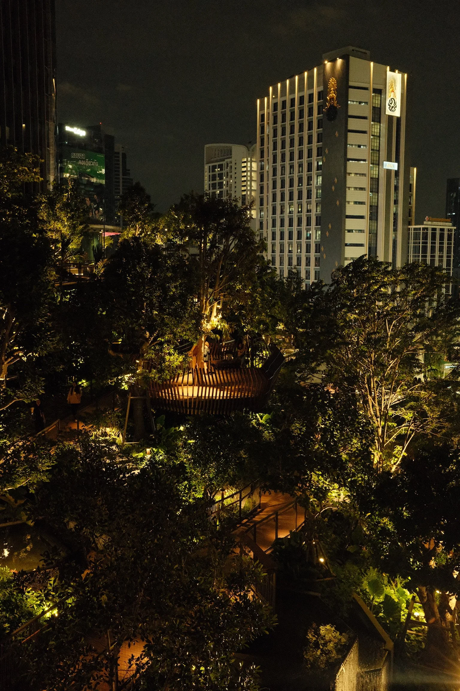 Een avondfoto van een moderne stad met hoge gebouwen en een groen park met verlichte wandelpaden en een houten uitkijkplatform tussen de bomen.