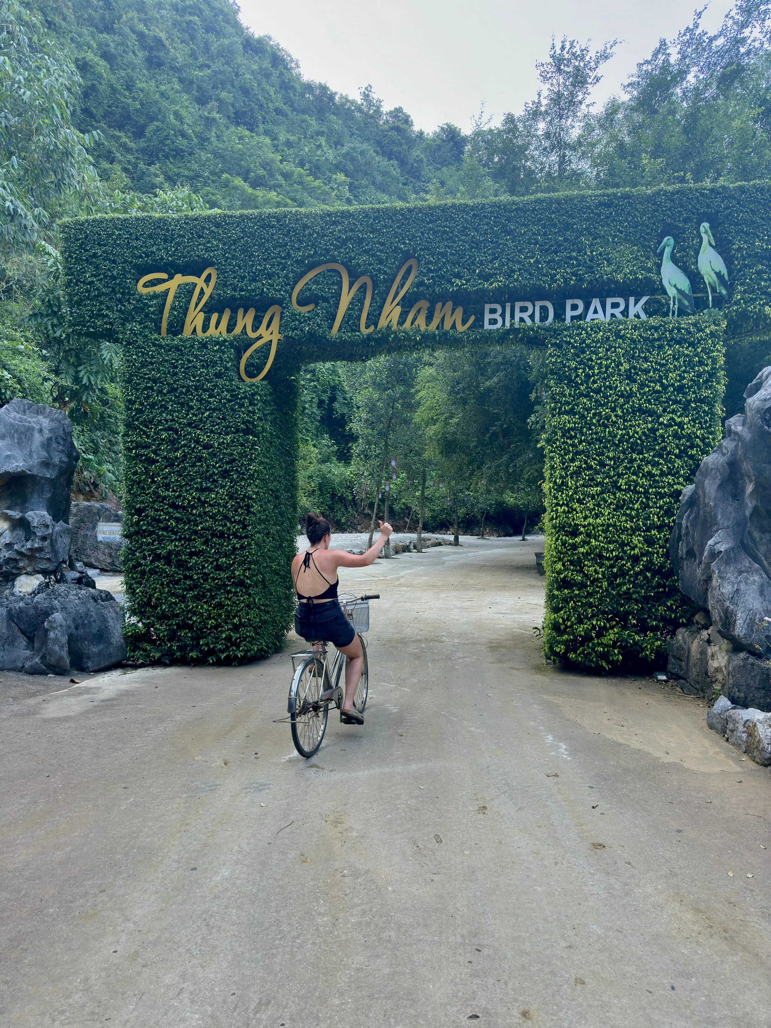 Toegangshek met de tekst 'Thung Nham Bird Park' en twee vogels op de bovenkant, omgeven door groene planten en rotsen, met een vrouw die op een fietst rijdt onder de poort.